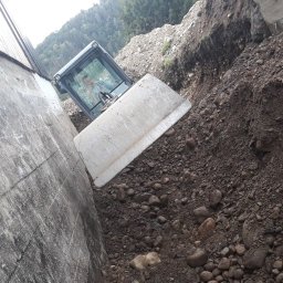 Michal Zolopa - Biała spycharka w wąskim wykopie, operator w kabinie. Ziemia z kamieniami. Betonowa ściana z boku. Praca na zboczu. Ujęcie z dołu.