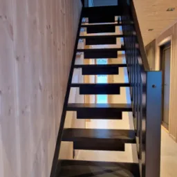 Nowoczesne, ażurowe schody z czarnymi stopniami i balustradą w jasnym wnętrzu z boazerią. Minimalistyczny design, perspektywa od dołu, widoczne oświetlenie.