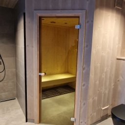 Michal Zolopa - Szklane drzwi do sauny z drewnianą obudową, widoczne drewniane ławki wewnątrz. Obok kabina prysznicowa i drewniana boazeria.