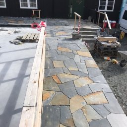 Michal Zolopa - Ścieżka z nieregularnych kamieni w odcieniach szarości i brązu prowadząca do domu z czarną fasadą i zielonymi drzwiami. Widoczne narzędzia i materiały budowlane.