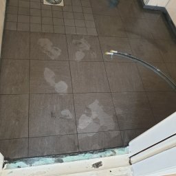 Michal Zolopa - Ułożone, ciemne płytki podłogowe w łazience, z widocznymi śladami wilgoci i rurami odpływowymi. Fragment progu drzwi.