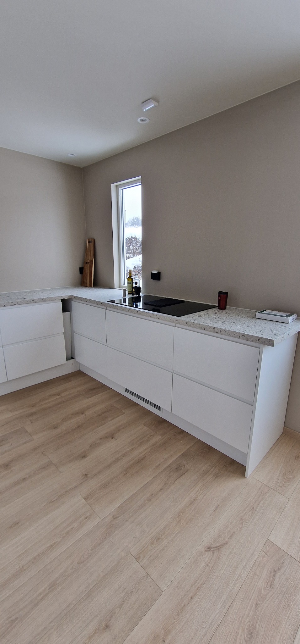 Nowoczesna, jasna kuchnia z białymi szafkami i blatem w plamki. Widoczna płyta indukcyjna, drewniana podłoga i okno z widokiem na zimowy krajobraz.