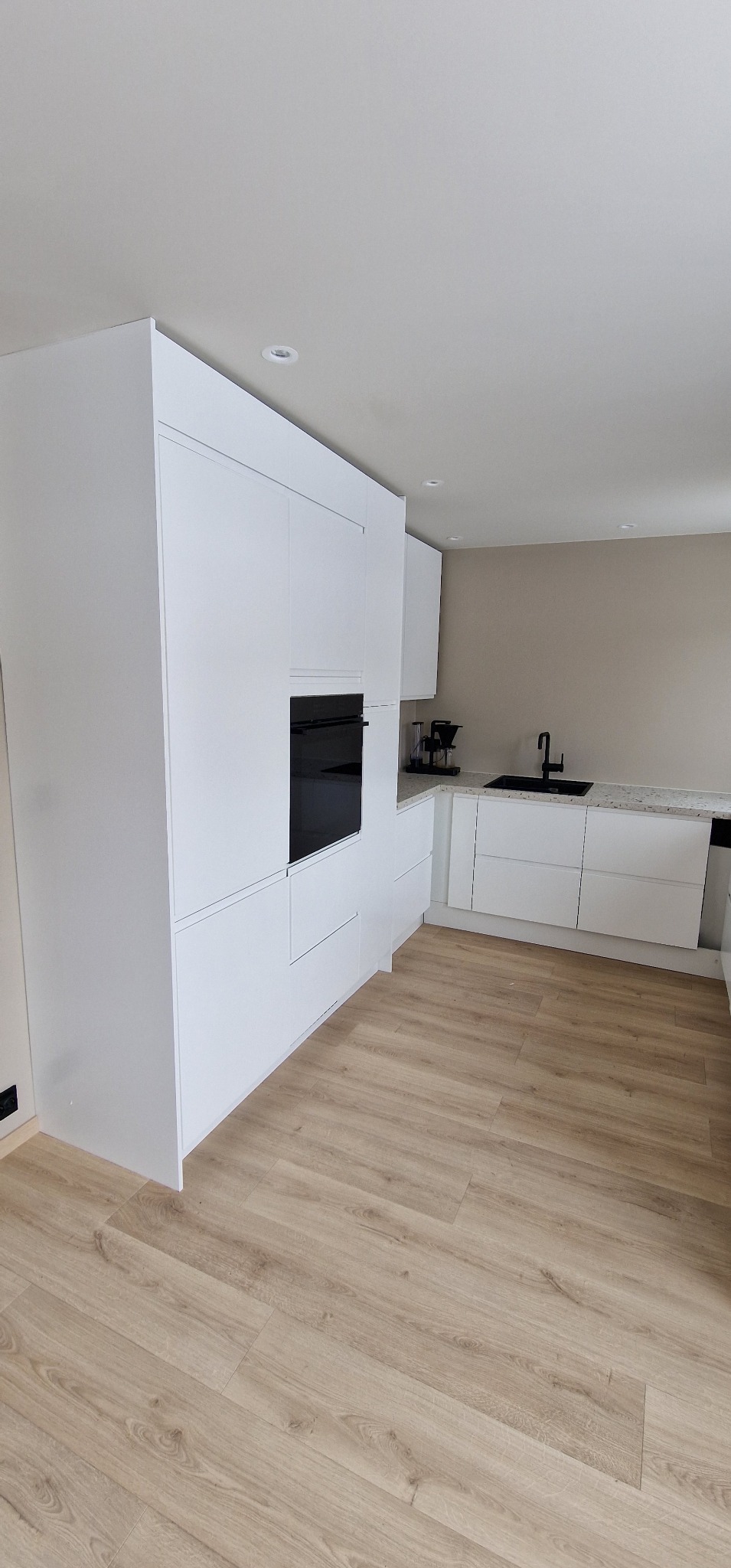 Minimalistyczna, biała kuchnia z zabudową po sufit i czarnymi akcentami armatury. Jasna podłoga z paneli imitujących drewno. Widok na zlewozmywak i ekspres do kawy.