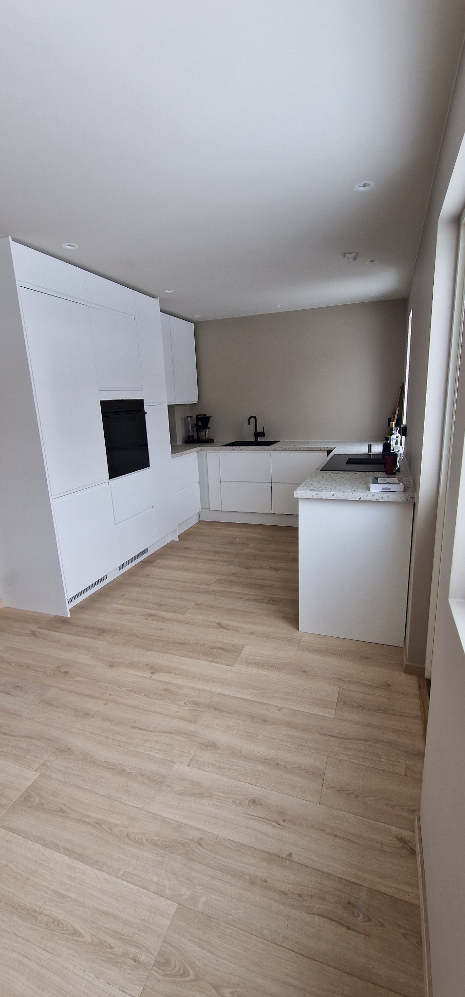 Nowoczesna, minimalistyczna kuchnia z białymi szafkami, czarnym piekarnikiem i drewnianą podłogą. Uwagę przyciąga czarna bateria zlewozmywakowa.