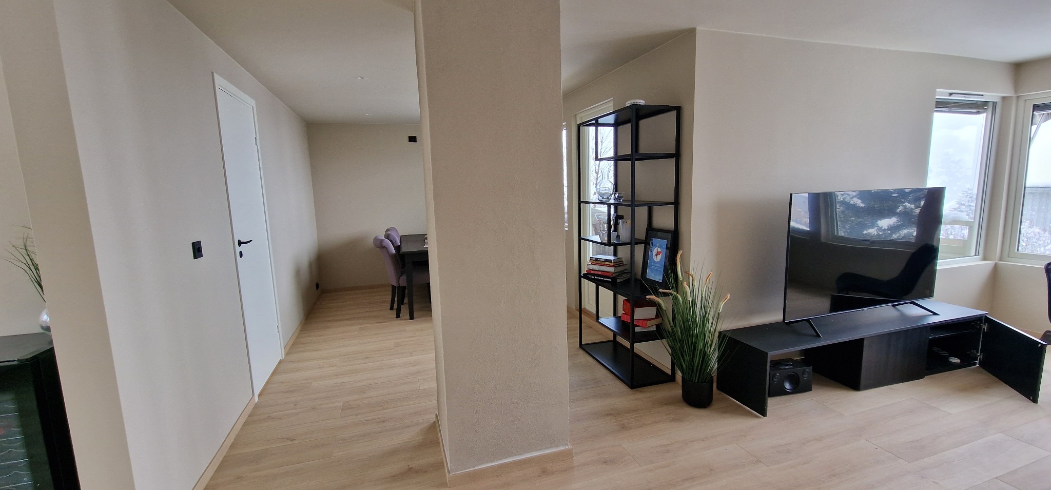 Jasne wnętrze po odświeżeniu: beżowe ściany, drewniana podłoga, czarne meble RTV i regał. W tle widoczny stół z fioletowymi krzesłami.