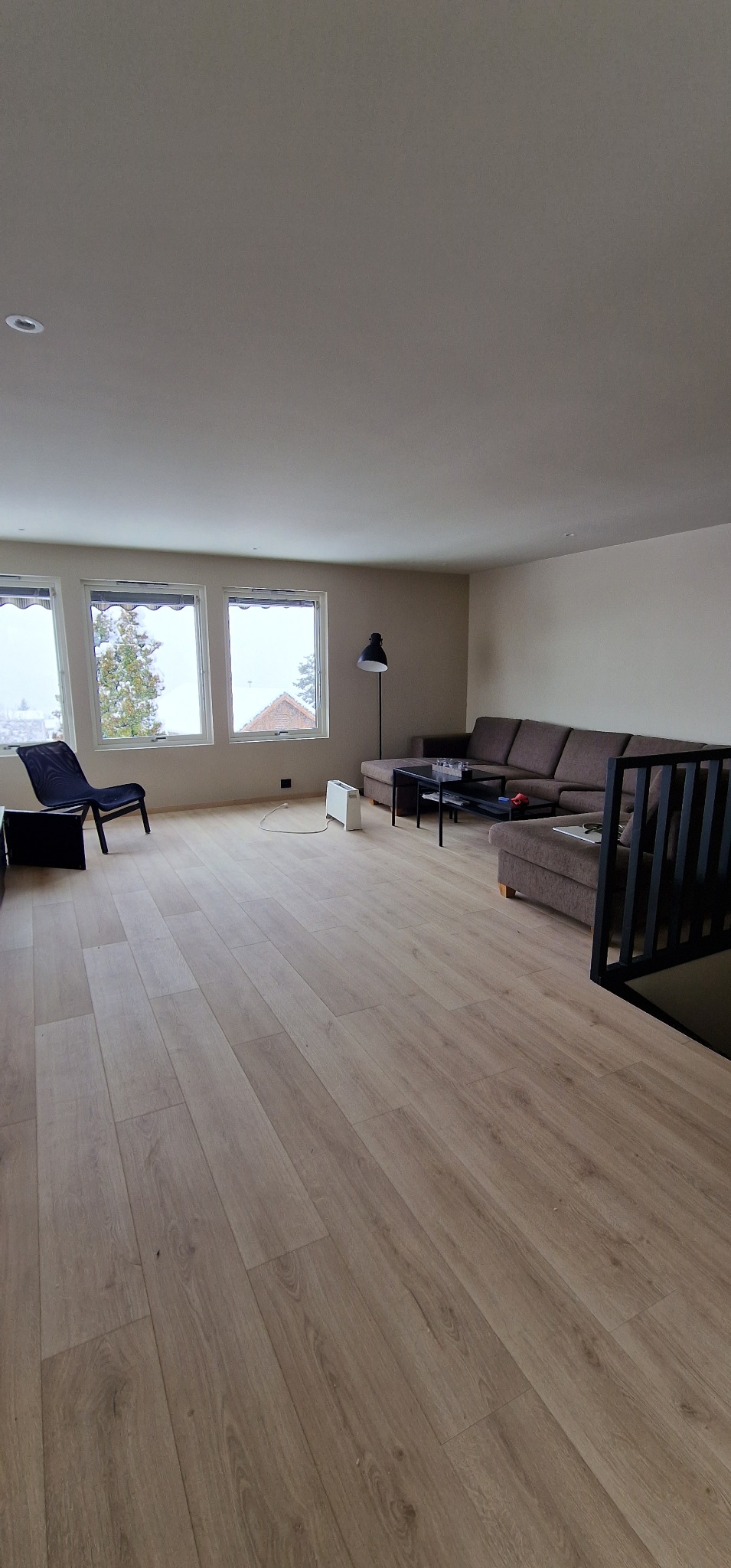 Jasna podłoga z paneli w przestronnym salonie z oknami i brązową sofą. Widoczny fragment czarnej balustrady schodów i elementy wyposażenia.