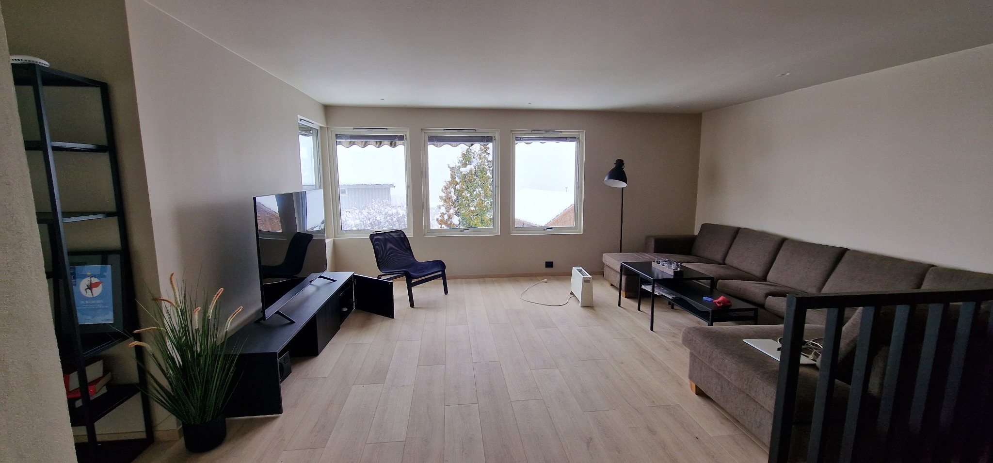 Jasne wnętrze salonu z parkietem, szarą sofą i czarnymi meblami, w tle widok na zaśnieżone drzewa przez okna. Minimalistyczny design, neutralna kolorystyka.