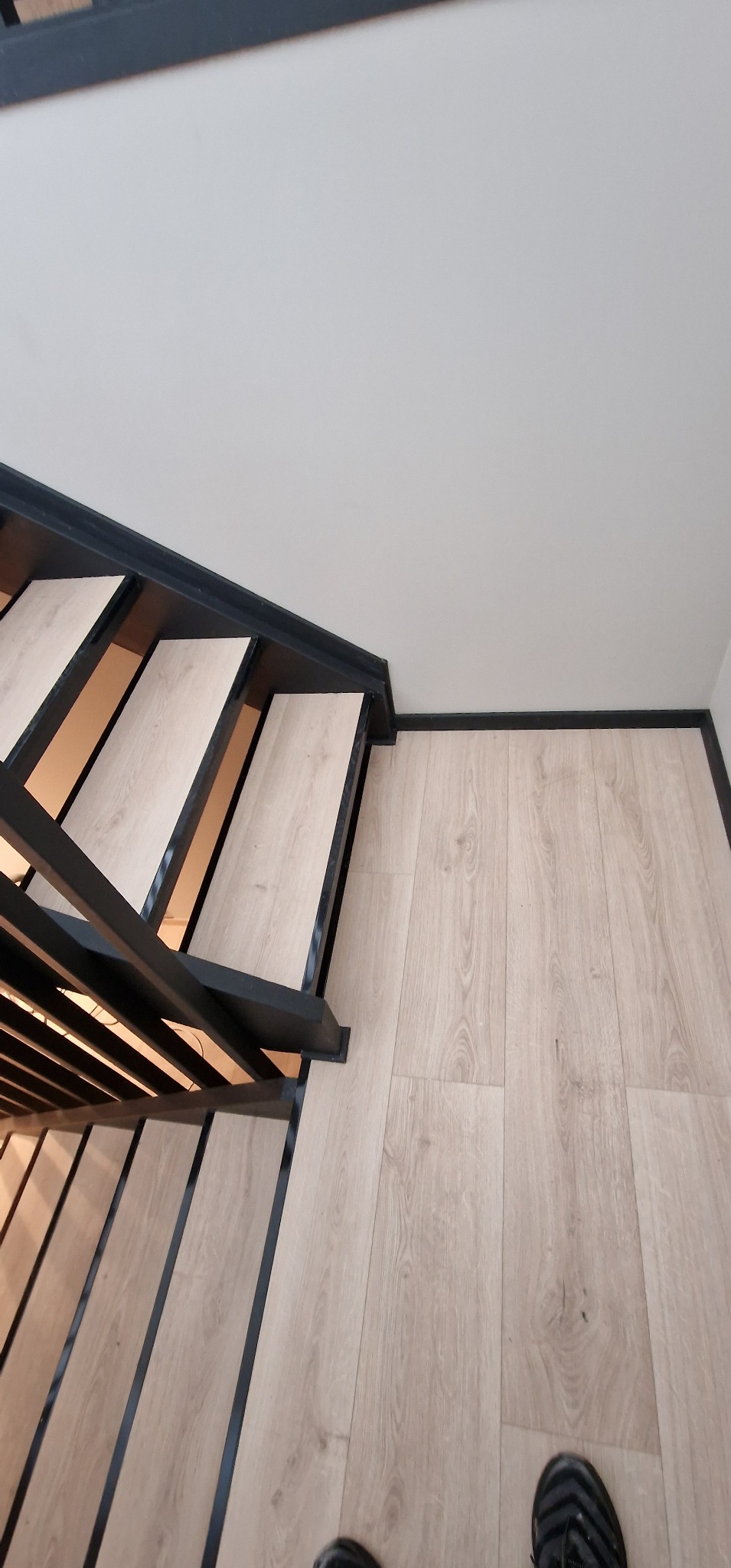 Nowoczesne schody z drewnianymi stopniami i czarną metalową balustradą, dopasowane do jasnej podłogi. Minimalistyczny design i precyzyjne wykończenie.