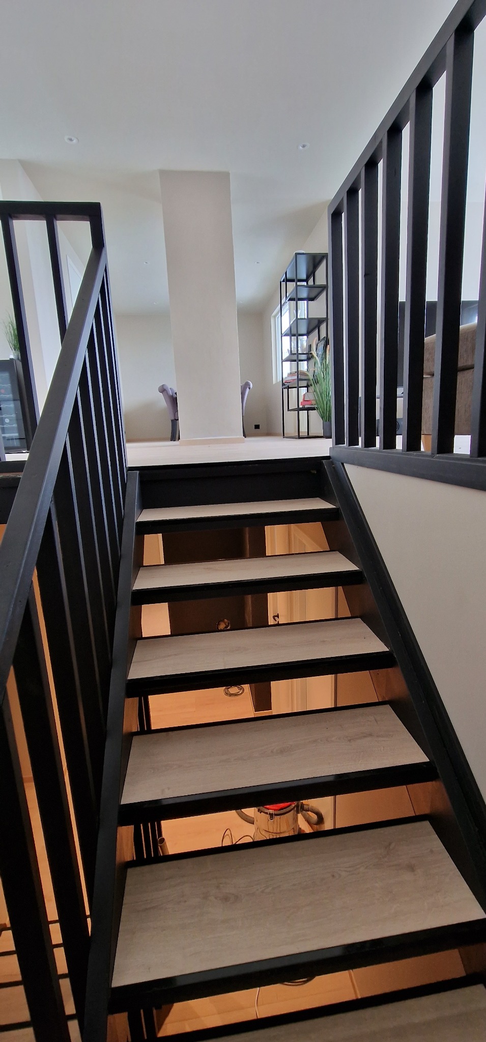 Schody wewnętrzne z drewnianymi stopniami i czarną balustradą, widziane z dołu. W tle jasne wnętrze z regałem i krzesłami.