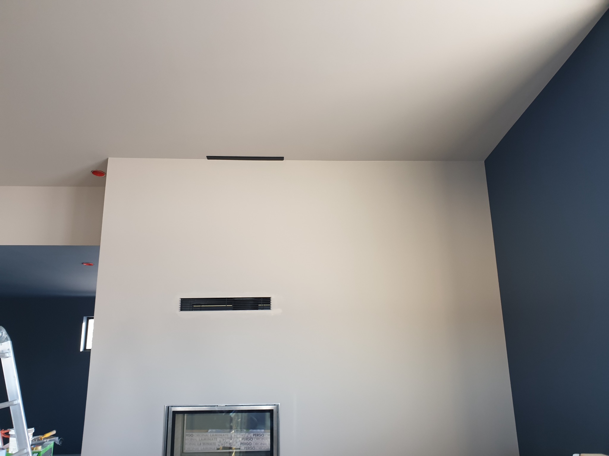 Wnętrze po szpachlowaniu i malowaniu: gładka, jasna ściana z kratkami wentylacyjnymi, fragment ściany w kolorze granatowym, drabina malarska w rogu.
