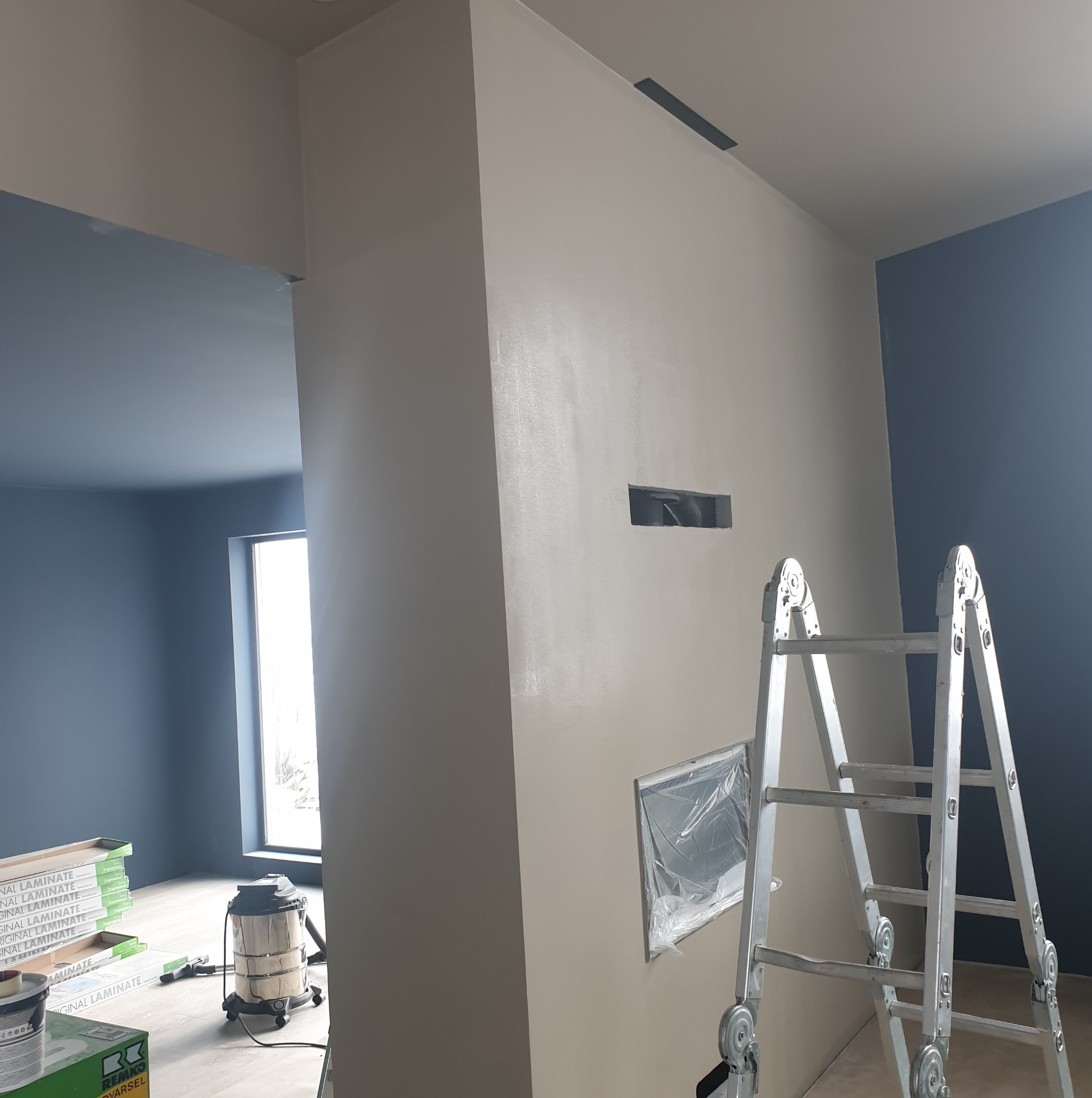 Świeżo wyszpachlowana ściana w beżowym odcieniu, widoczna drabina, zakryte okno folią, podłoga częściowo zakryta, przygotowana do dalszych prac.