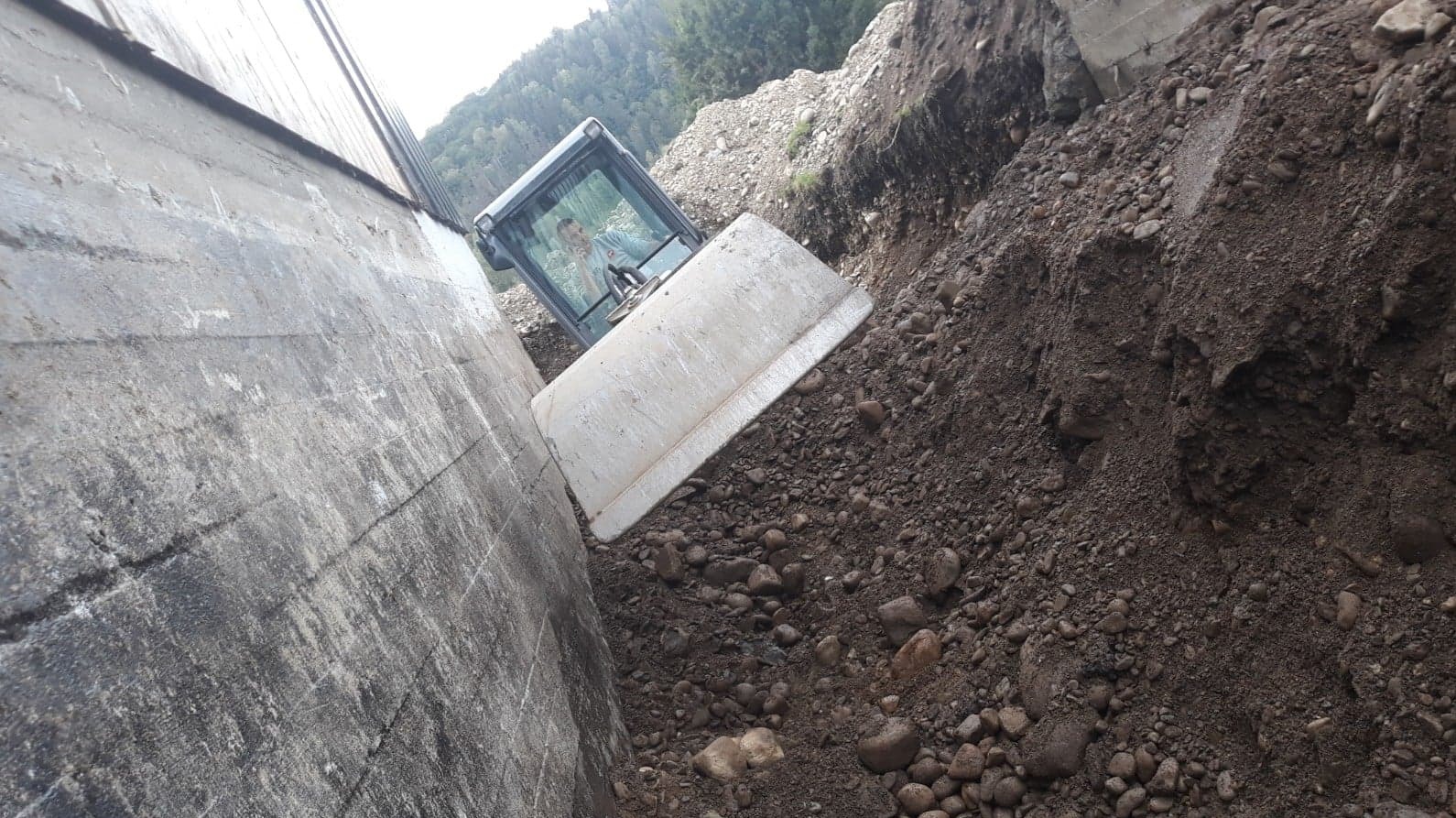 Biała spycharka w wąskim wykopie, operator w kabinie. Ziemia z kamieniami. Betonowa ściana z boku. Praca na zboczu. Ujęcie z dołu.