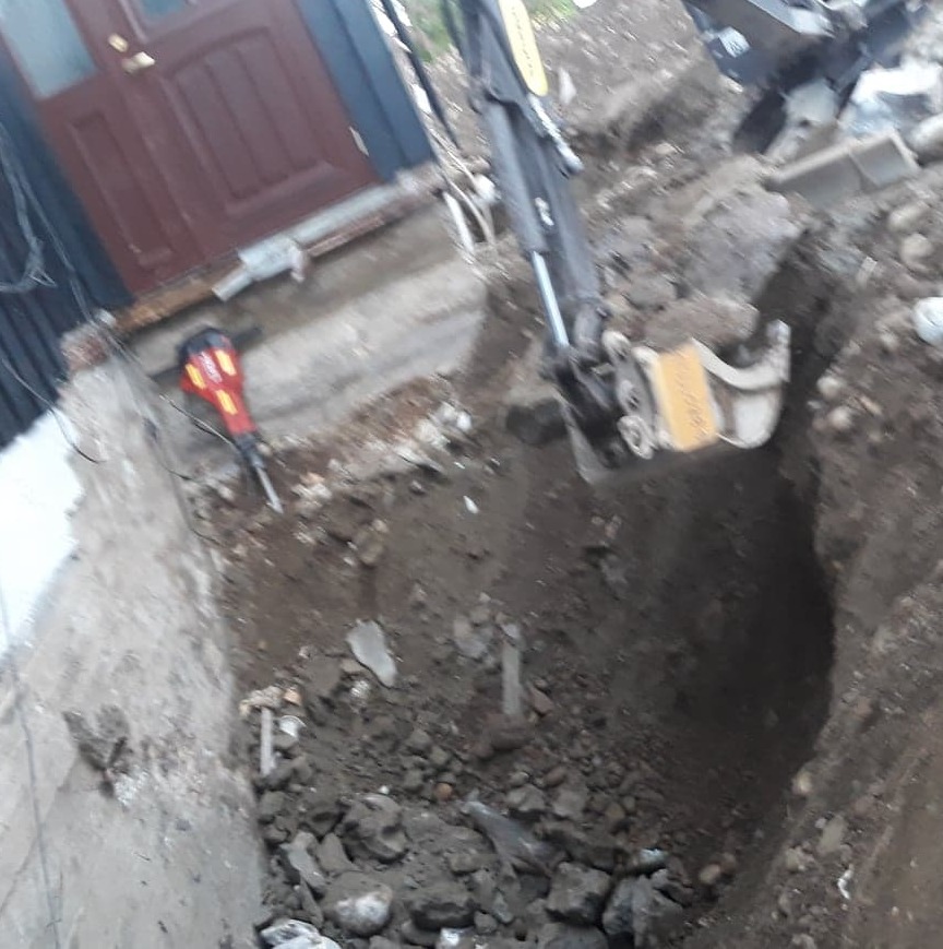 Wykop pod fundament z widoczną koparką i młotem pneumatycznym przy wejściu do budynku. Prace ziemne w toku, gruz i ziemia w wykopie.