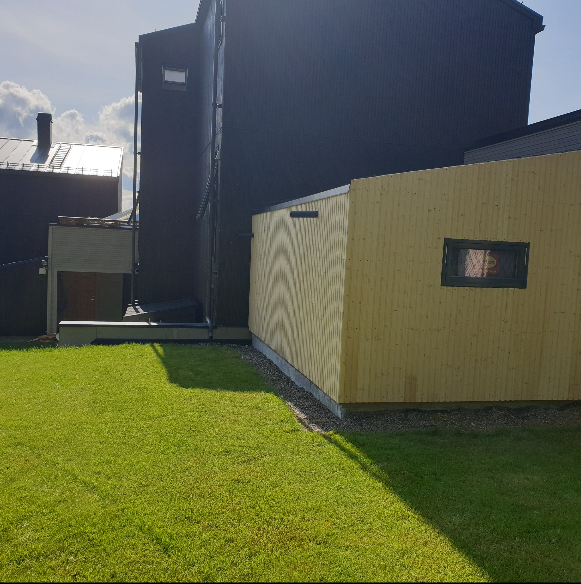 Nowa, jasna, drewniana konstrukcja z małym oknem, przylegająca do ciemnego budynku na tle zielonego trawnika w słoneczny dzień.