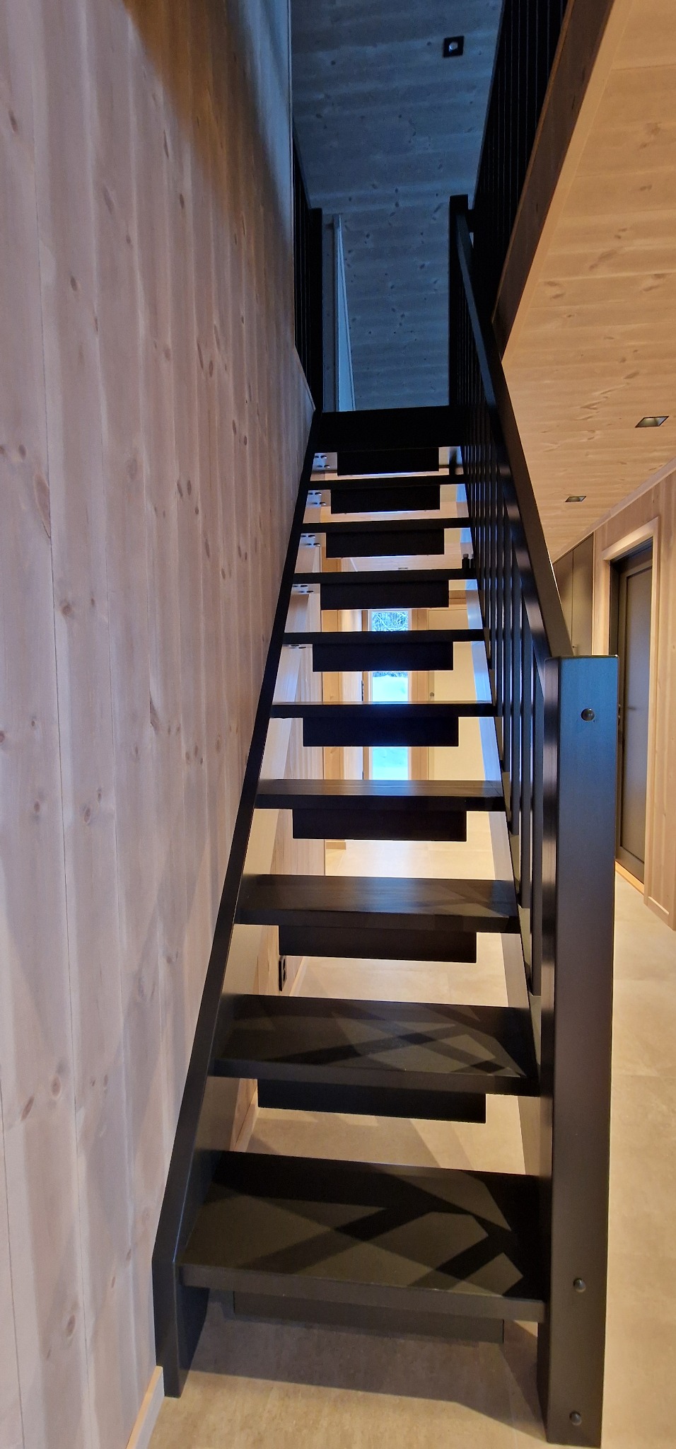 Nowoczesne, ażurowe schody z czarnymi stopniami i balustradą w jasnym wnętrzu z boazerią. Minimalistyczny design, perspektywa od dołu, widoczne oświetlenie.