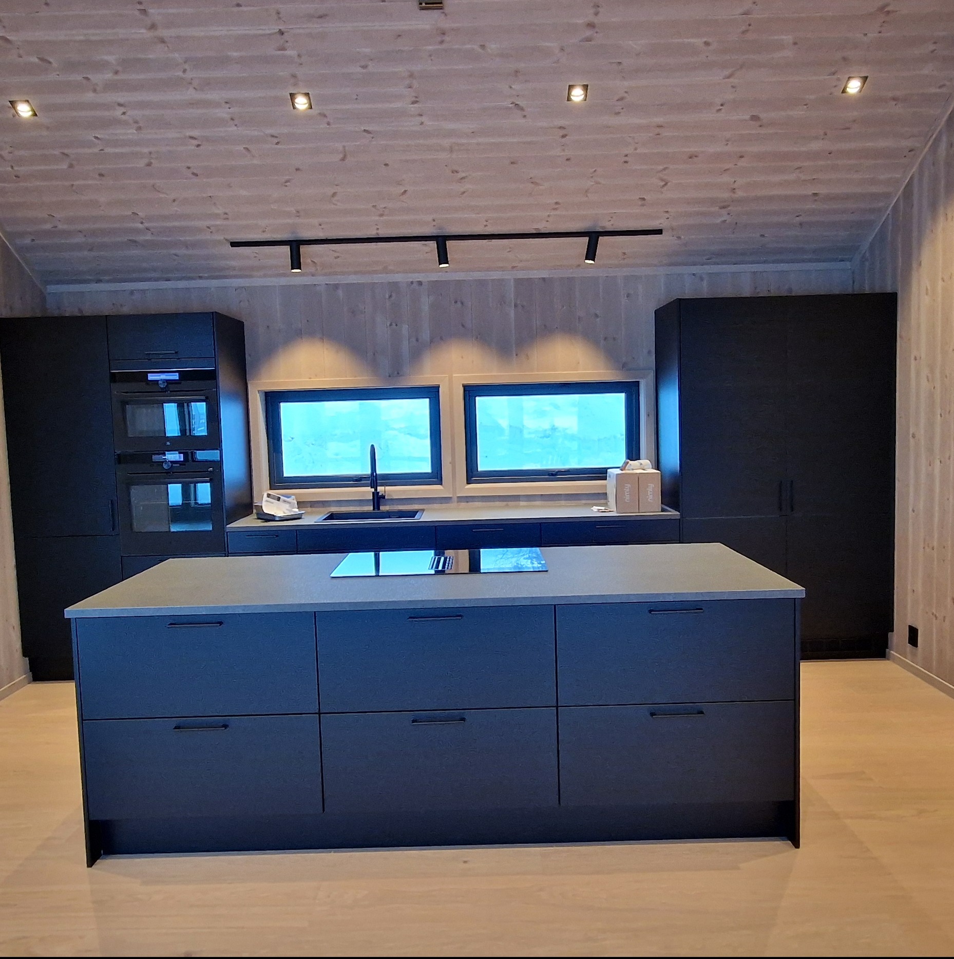 Nowoczesna kuchnia z wyspą i czarnymi szafkami, drewniane wykończenie ścian i sufitu, oświetlenie punktowe i okna z widokiem na zewnątrz. Minimalistyczny design.