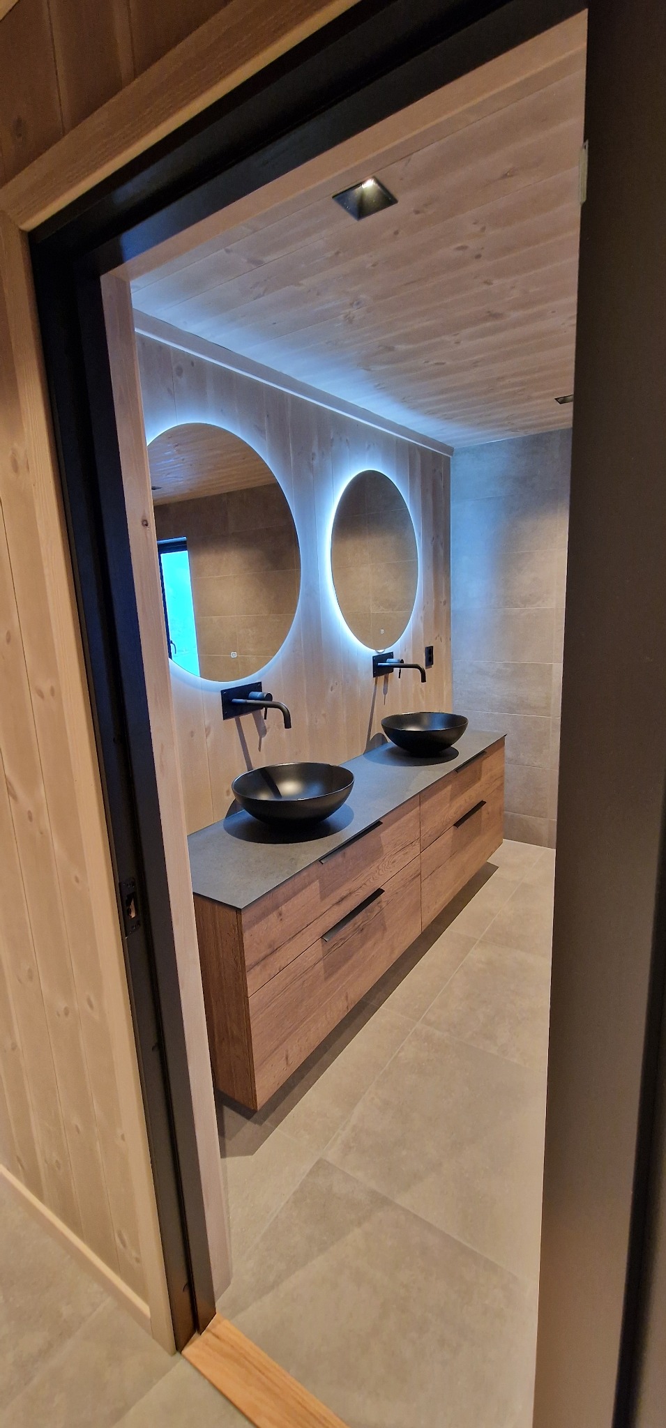 Nowoczesna łazienka z drewnianą szafką pod umywalki, okrągłe lustra z podświetleniem LED i czarne baterie, widok przez otwarte drzwi.