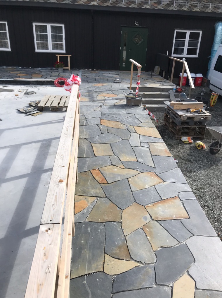 Ścieżka z nieregularnych kamieni w odcieniach szarości i brązu prowadząca do domu z czarną fasadą i zielonymi drzwiami. Widoczne narzędzia i materiały budowlane.