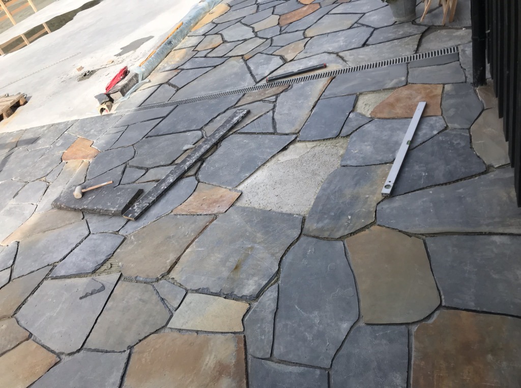 Układanie nieregularnej, kamiennej posadzki. Fragment ułożonej nawierzchni z narzędziami: poziomica, młotek i listwa, widoczny drenaż liniowy.