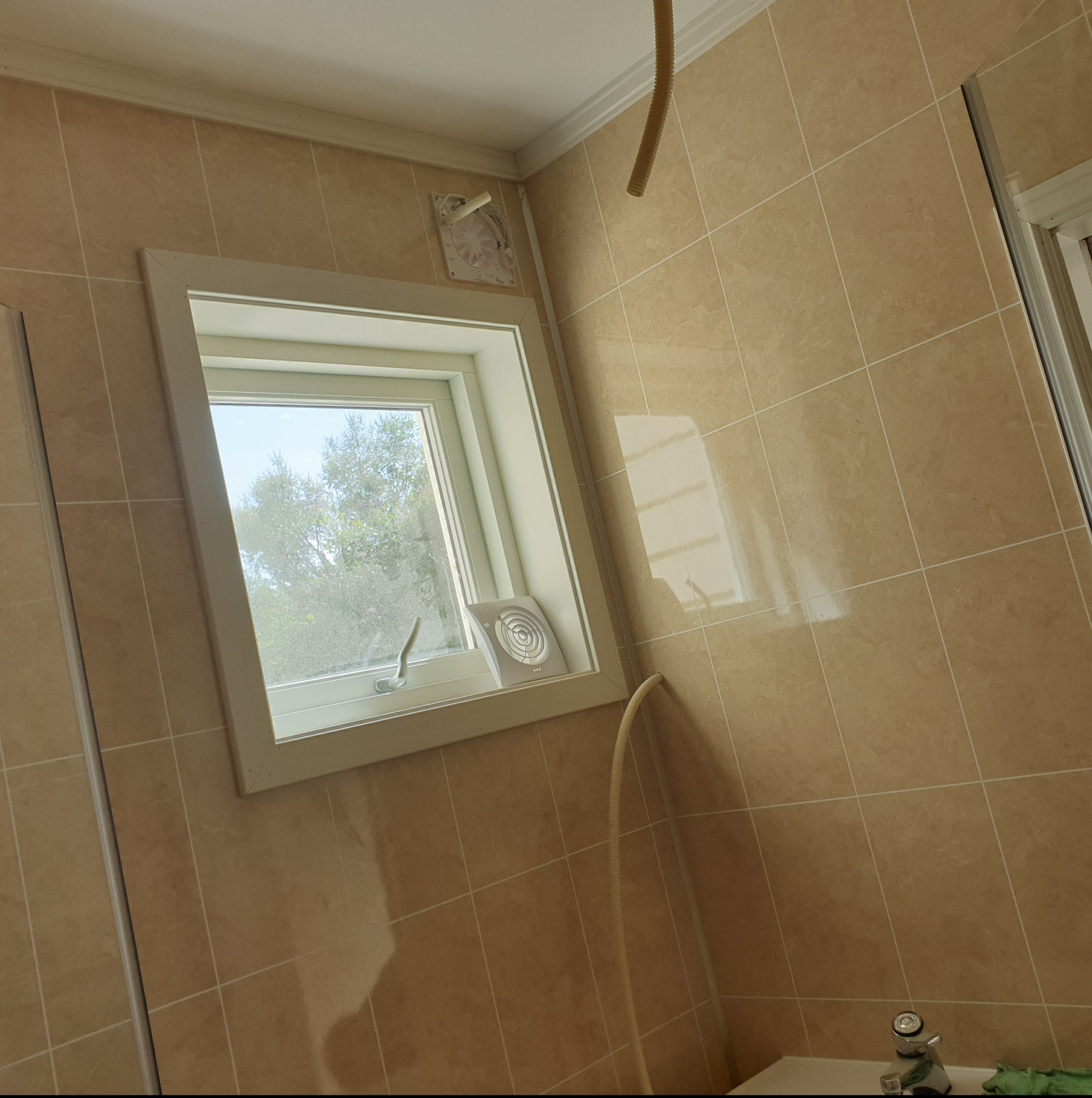 Beżowe płytki w łazience z oknem, wentylatorem i fragmentem rury wentylacyjnej. Widok na zewnątrz przez okno.