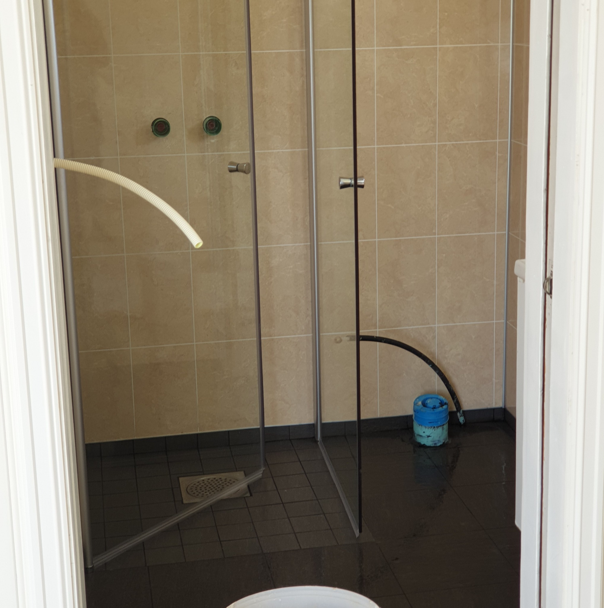 Kabina prysznicowa z brązowymi płytkami, widoczne rury i niebieskie wiadro. Ujęcie zza białych futryn drzwi, ukazujące etap prac w łazience.