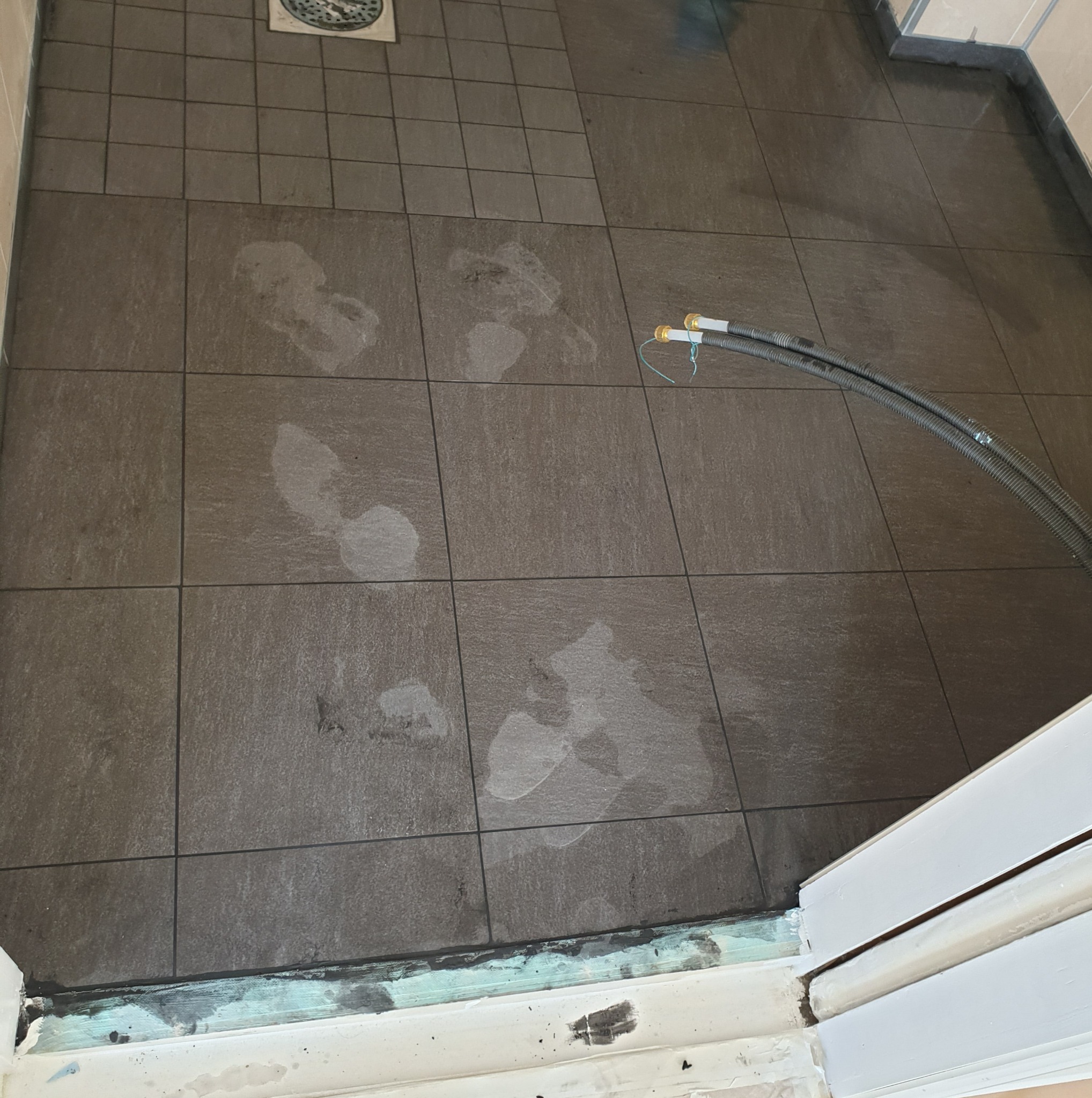 Ułożone, ciemne płytki podłogowe w łazience, z widocznymi śladami wilgoci i rurami odpływowymi. Fragment progu drzwi.