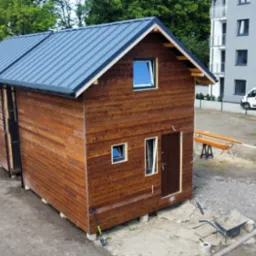 Brązowy, drewniany dom modułowy z szarym dachem na fundamencie betonowym. Na zewnątrz widoczny wózek, drabina i narzędzia. W tle budynek mieszkalny i samochody dostawcze.