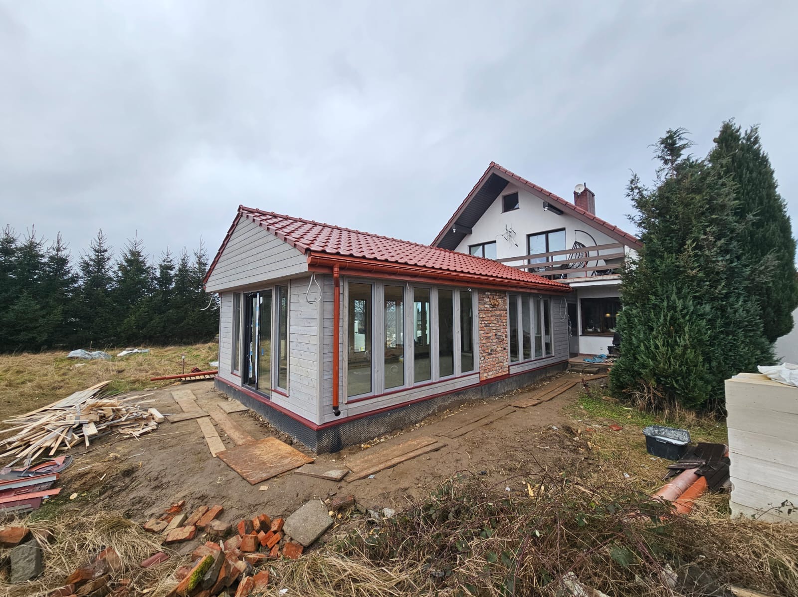 Do domu dobudowana jasna, przeszklona konstrukcja z czerwonym dachem. Widoczne materiały budowlane i otoczenie w trakcie prac wykończeniowych.