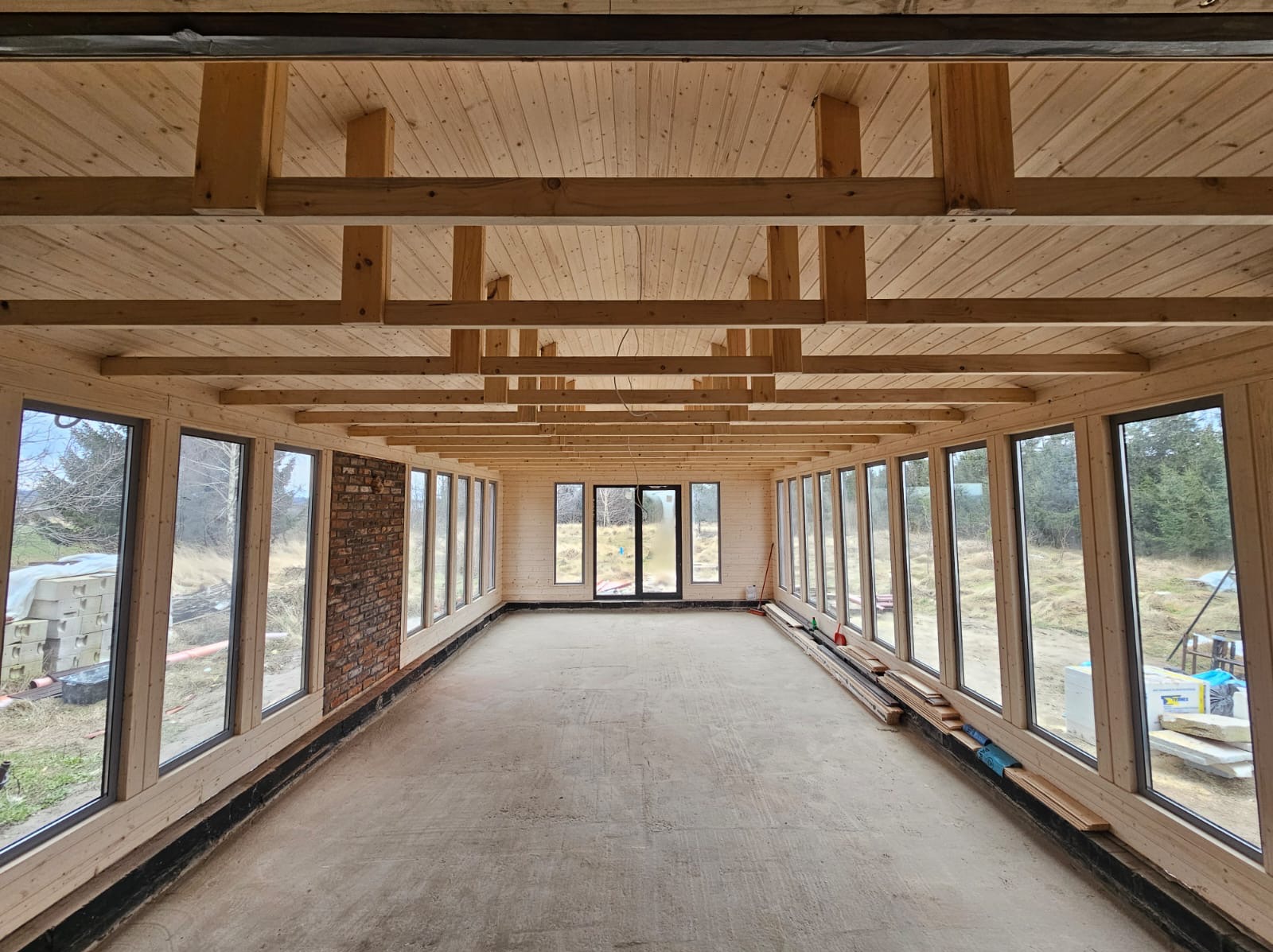 Wnętrze surowego domu szkieletowego z drewnianą konstrukcją stropu, dużymi oknami i fragmentem ściany z cegły. Widoczny stan deweloperski z surową podłogą.