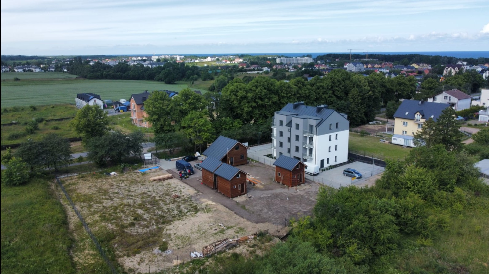 Widok z drona na plac budowy z trzema małymi domkami z ciemnego drewna i budynkiem mieszkalnym w tle. W tle widoczne miasto i morze.