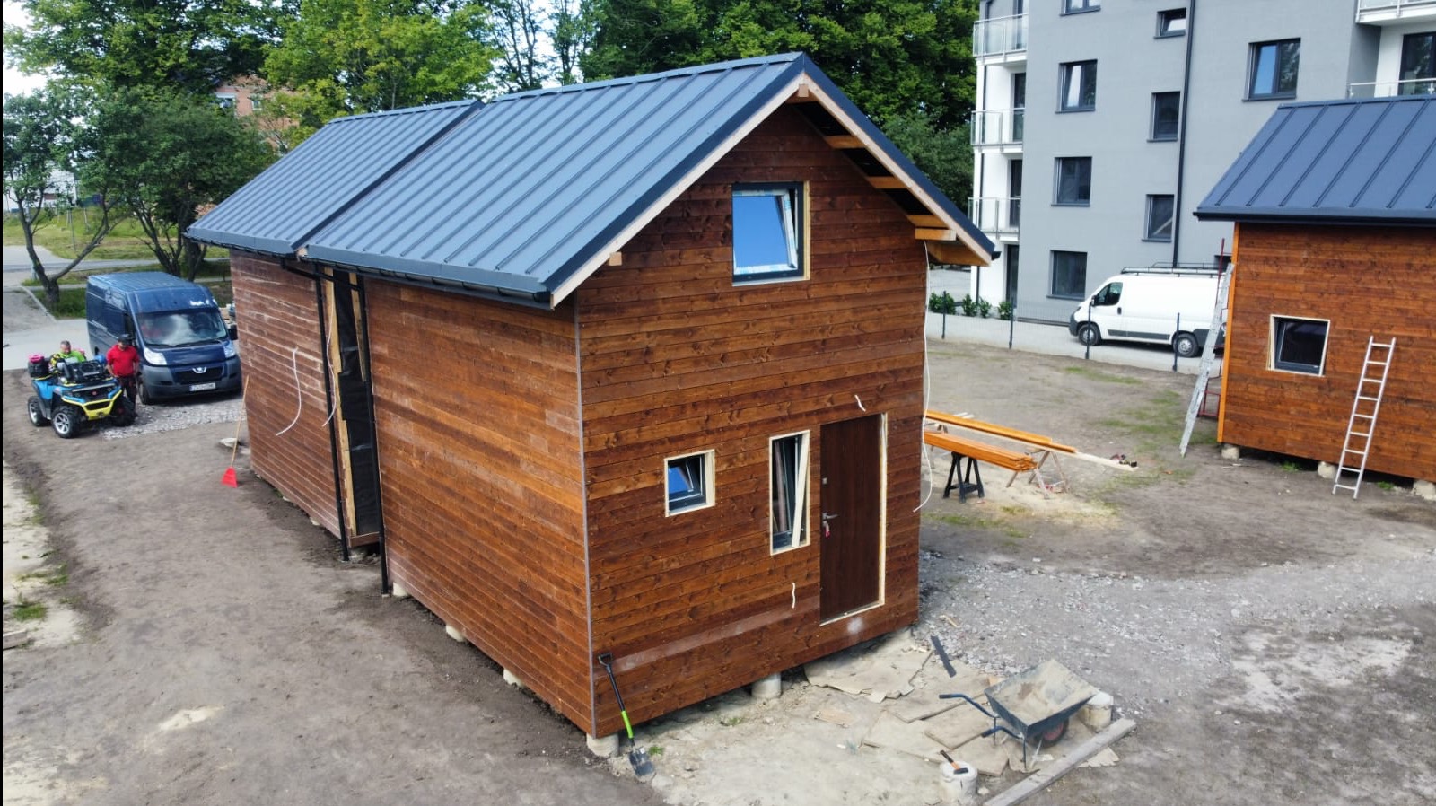 Brązowy, drewniany dom modułowy z szarym dachem na fundamencie betonowym. Na zewnątrz widoczny wózek, drabina i narzędzia. W tle budynek mieszkalny i samochody dostawcze.
