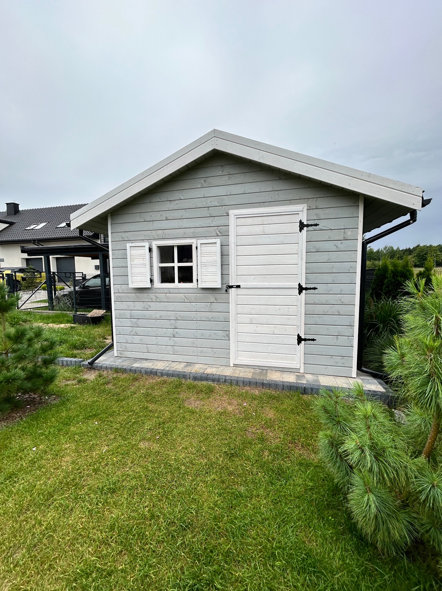 Drewniany domek narzędziowy w odcieniach szarości z białymi detalami (okiennice, drzwi) na tle zielonego trawnika i ogrodzenia. Widoczny fragment domu w tle.