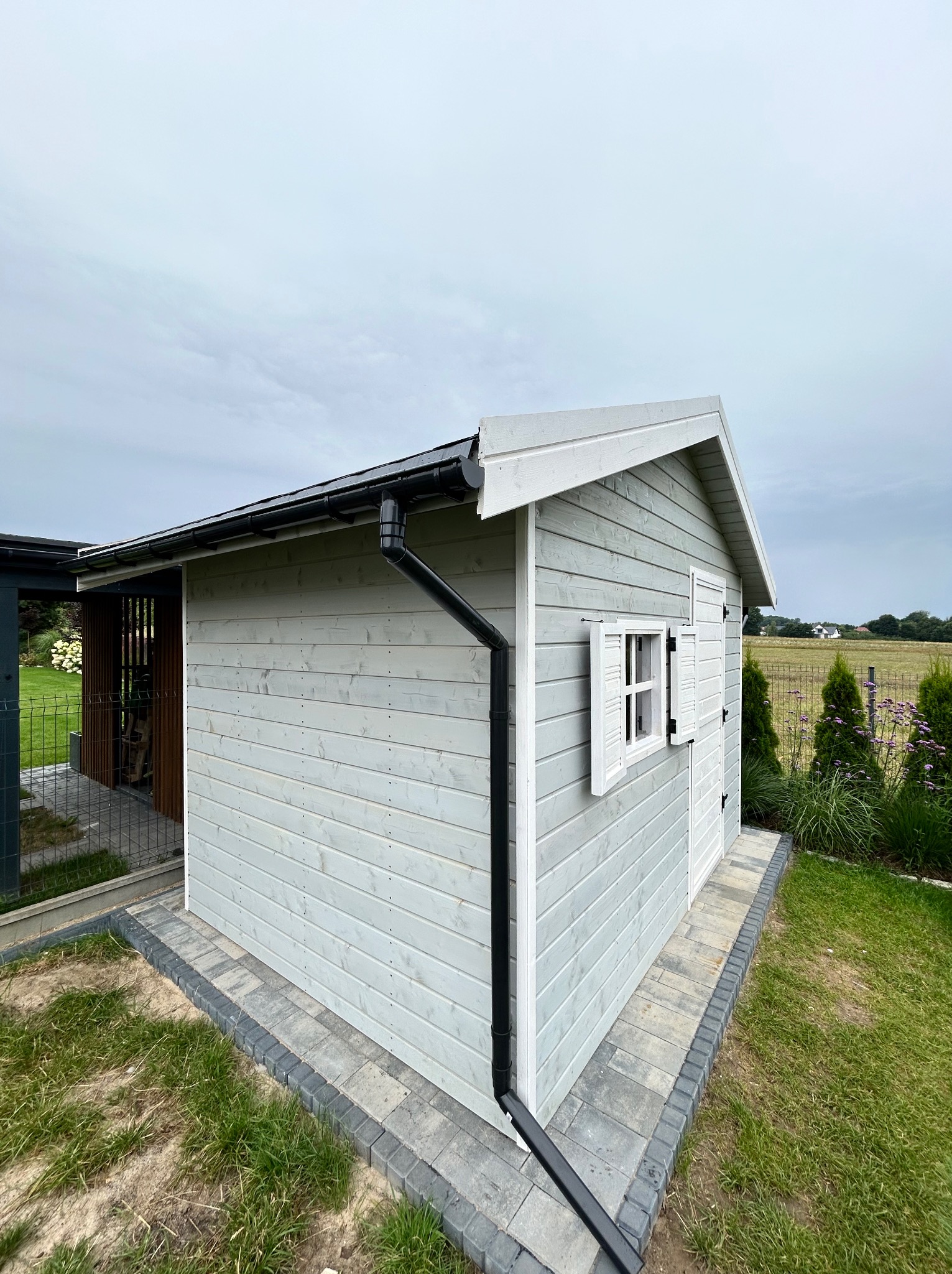 Drewniana altana w szarym kolorze z białymi okiennicami i czarną rynną, otoczona zielonym trawnikiem i kostką brukową.