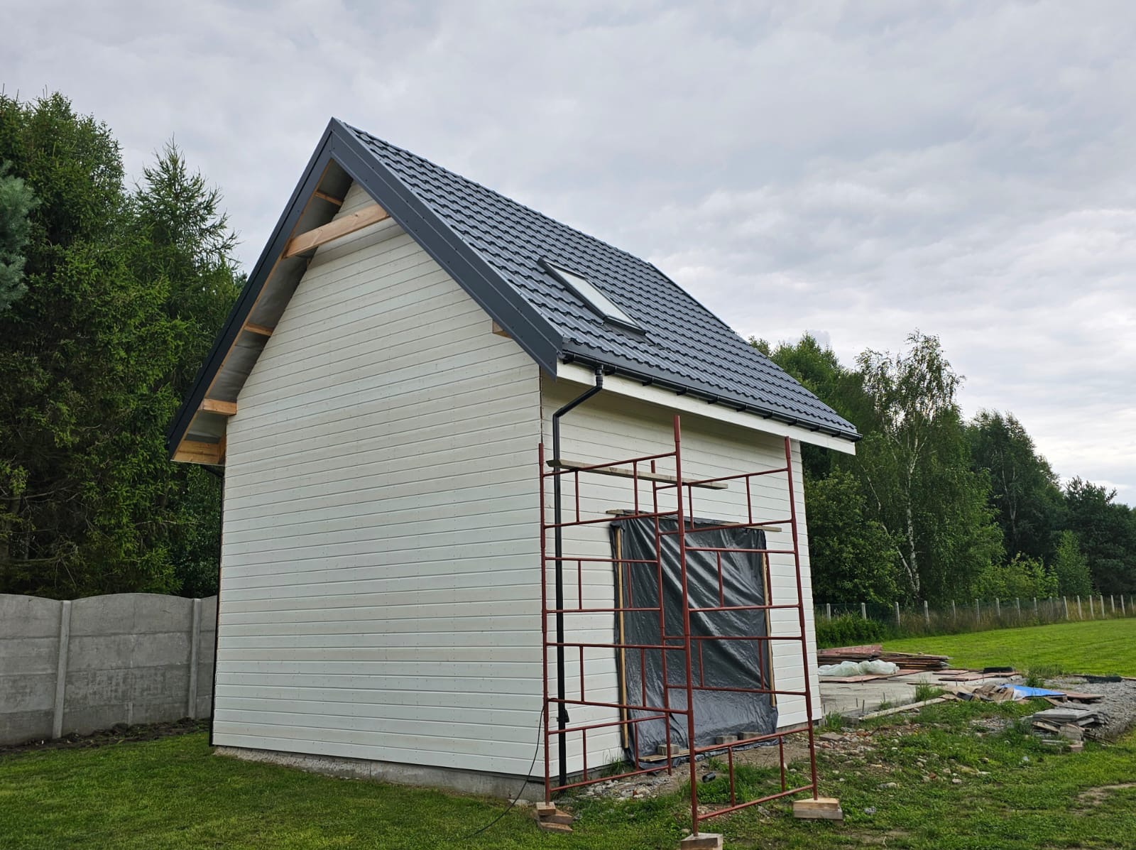 Niedokończony, parterowy dom szkieletowy z ciemnym dachem i oknem dachowym. Ściany pokryte jasną, poziomą boazerią. Widoczne rusztowanie przykryte folią.