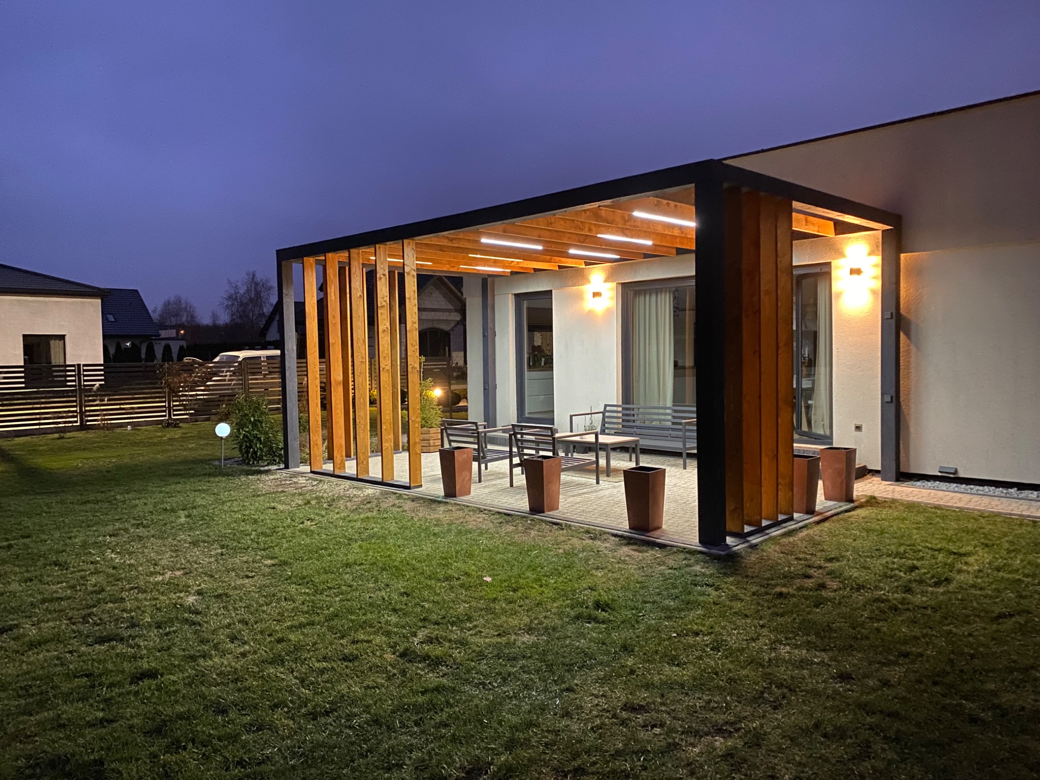 Oświetlona pergola drewniana z elementami czarnymi, stojąca na tle domu z oknami, z meblami ogrodowymi i donicami na kostce brukowej. Wieczorny krajobraz.
