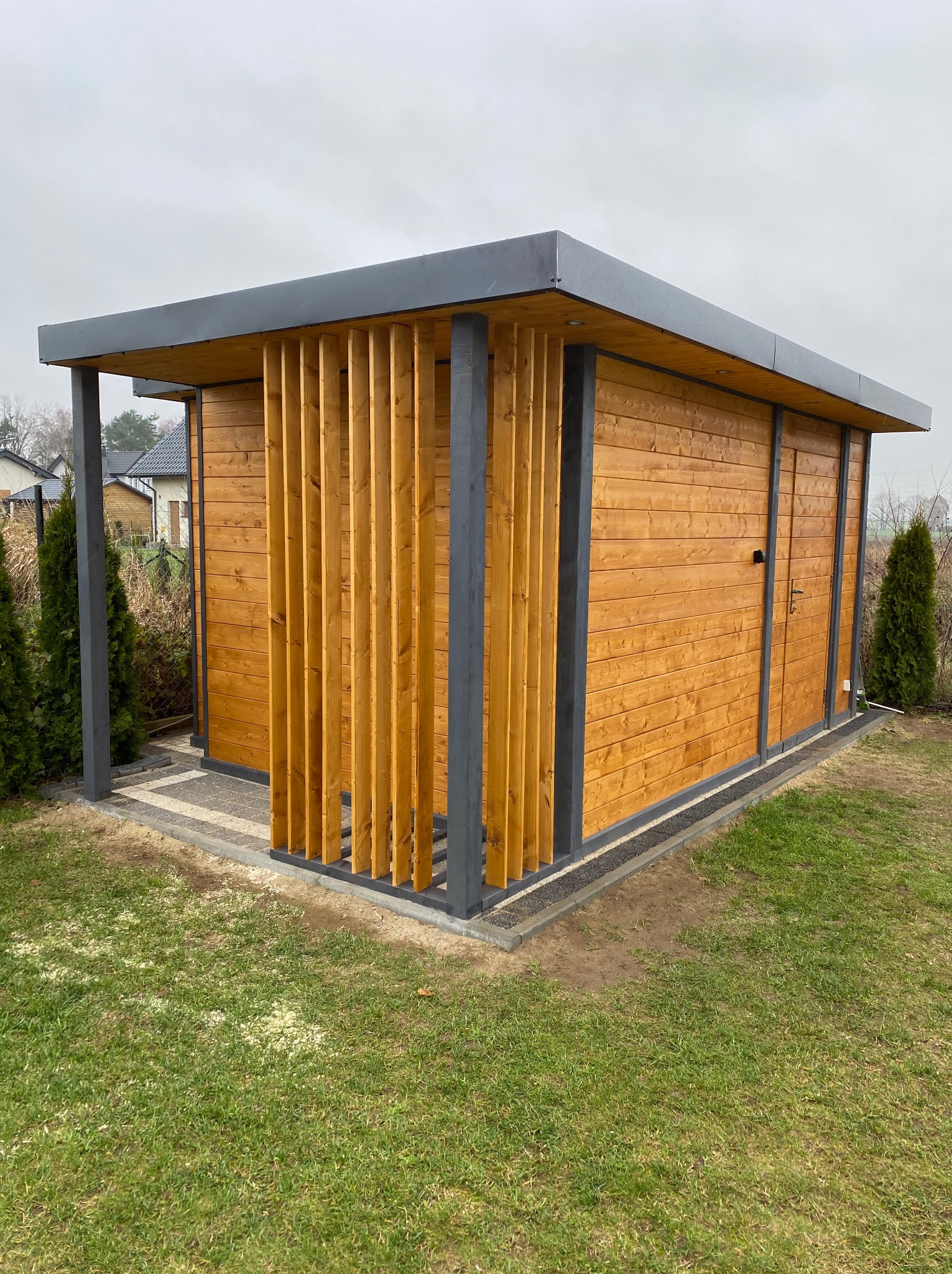 Nowoczesna, drewniana altana z szarymi elementami konstrukcyjnymi i pionowymi lamelami, otoczona zielonym trawnikiem. Widok z zewnątrz, w tle zabudowa jednorodzinna.