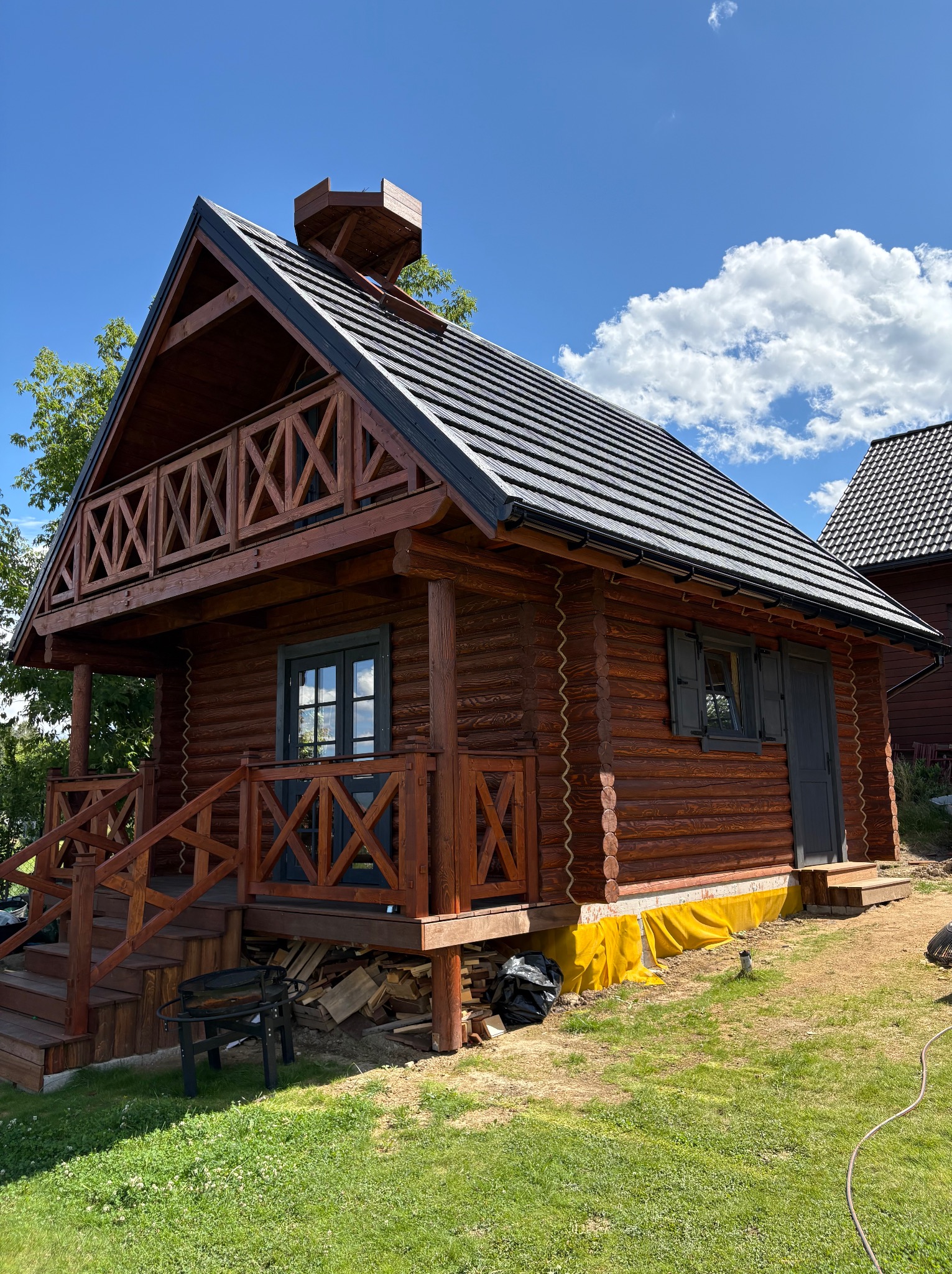 Drewniany domek letniskowy z balkonem i schodami, widoczny z zewnątrz w słoneczny dzień. Na trawie leżą drewno i grill. Fundament zabezpieczony żółtą folią.