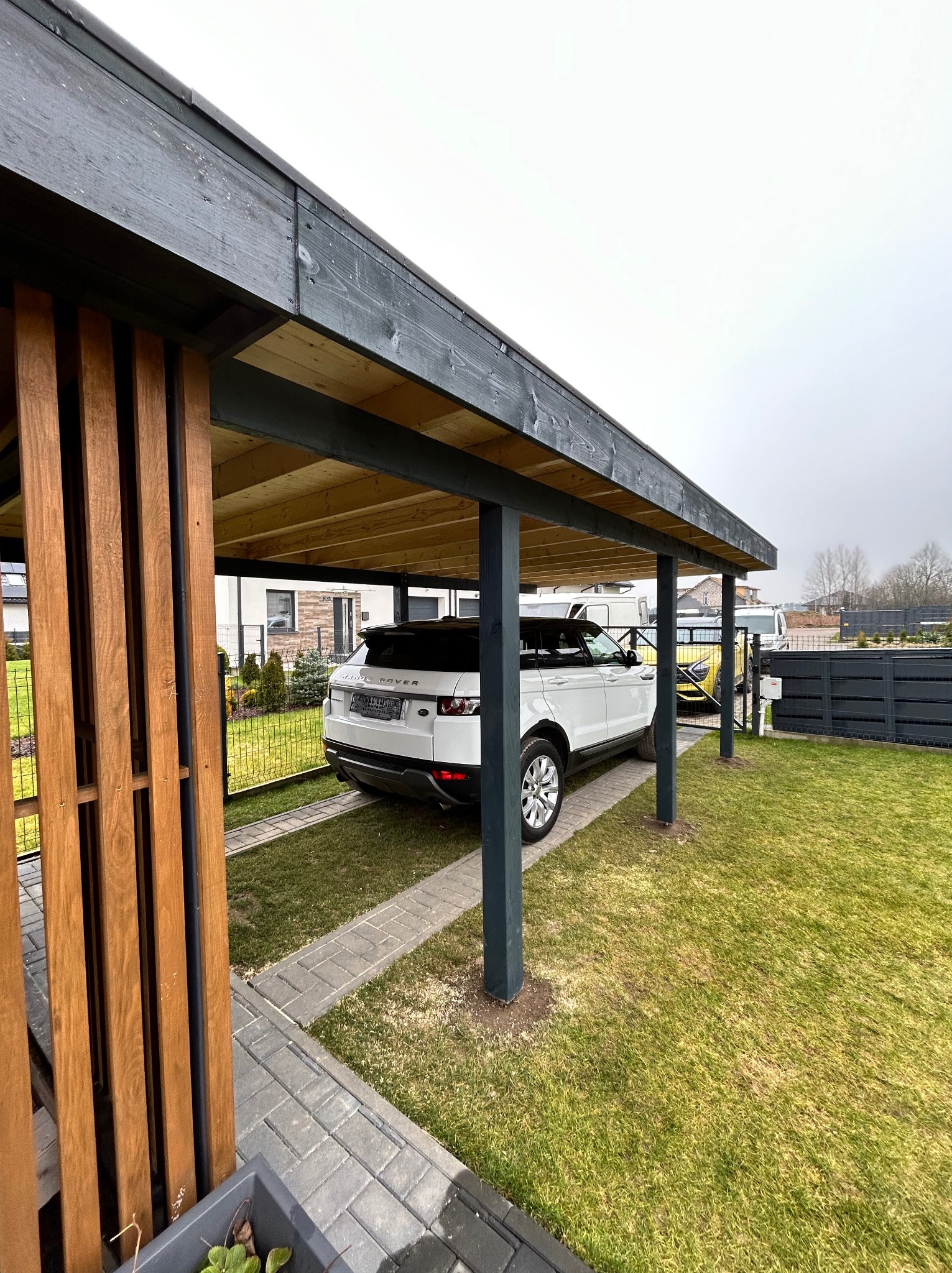 Drewniana wiata garażowa z ażurową ścianką boczną, kryjąca biały samochód Range Rover, na tle zadbanego trawnika i ogrodzenia panelowego.