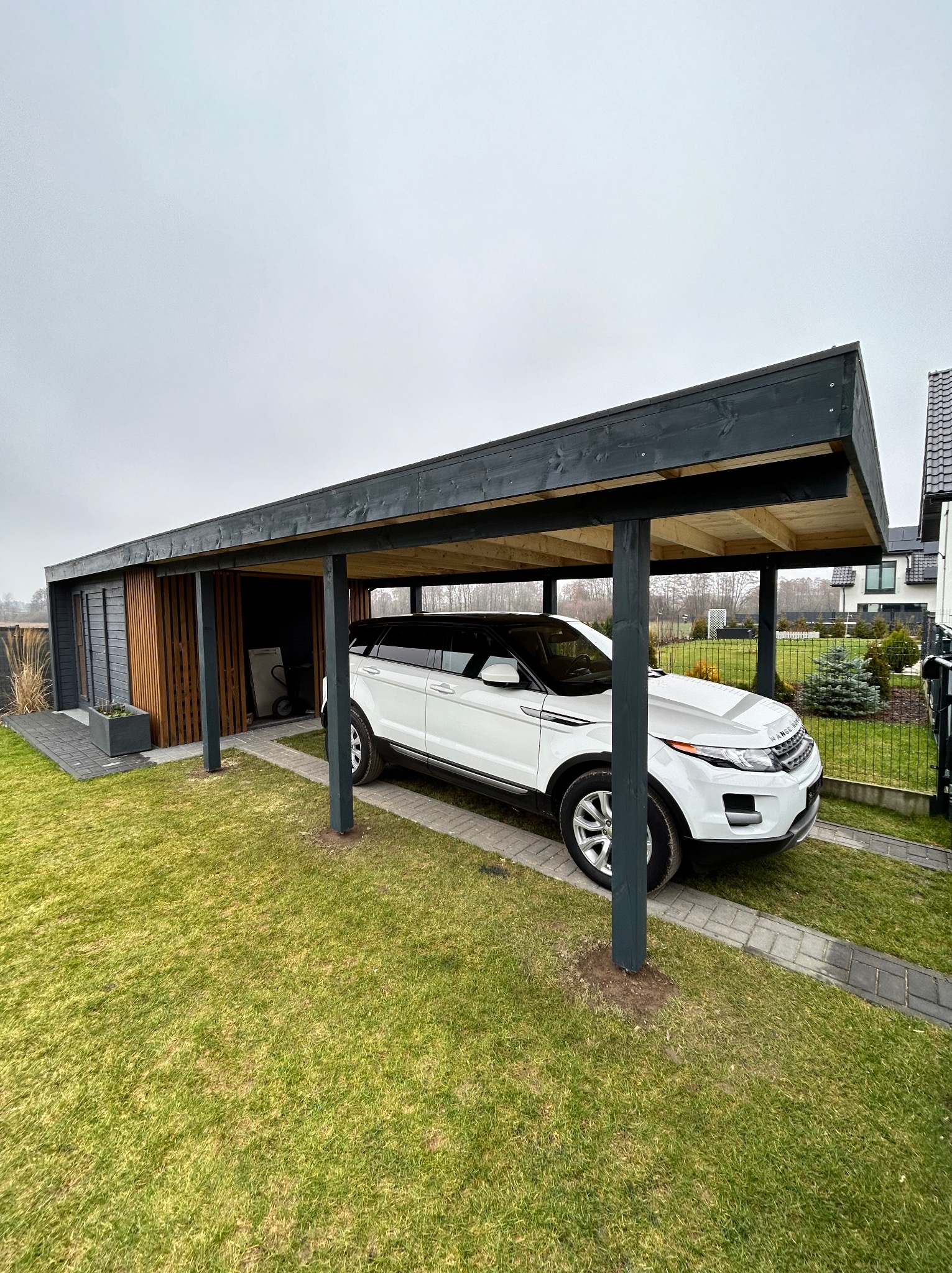 Nowoczesna, grafitowa wiata garażowa z drewna, z białym SUV-em w środku, na tle zadbanego trawnika i domu jednorodzinnego. Widoczne detale wykończenia i oświetlenie.