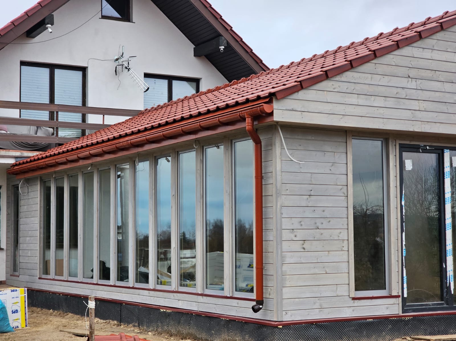 Jasna, drewniana dobudówka z dużymi oknami i brązowym orynnowaniem, przylegająca do białego domu z anteną na ścianie. Widoczne materiały budowlane.