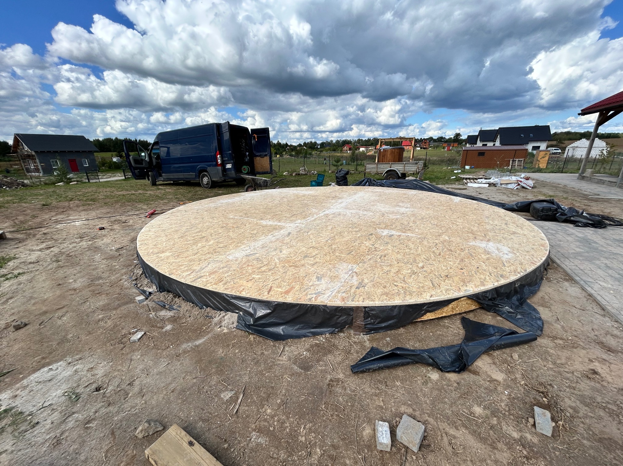 Okrągła platforma z płyty OSB, zabezpieczona folią, na placu budowy. W tle bus, domki i pochmurne niebo. Widoczny etap budowy konstrukcji.
