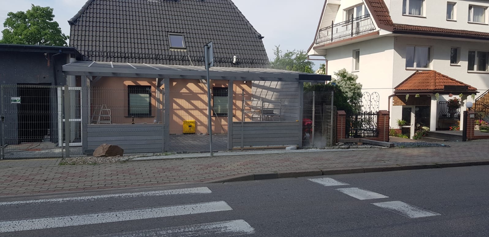 Zadaszenie tarasu z szarego drewna, widoczne z ulicy na tle dwóch domów. Metalowe ogrodzenie i drabina oparta o ścianę sugerują trwające prace wykończeniowe.