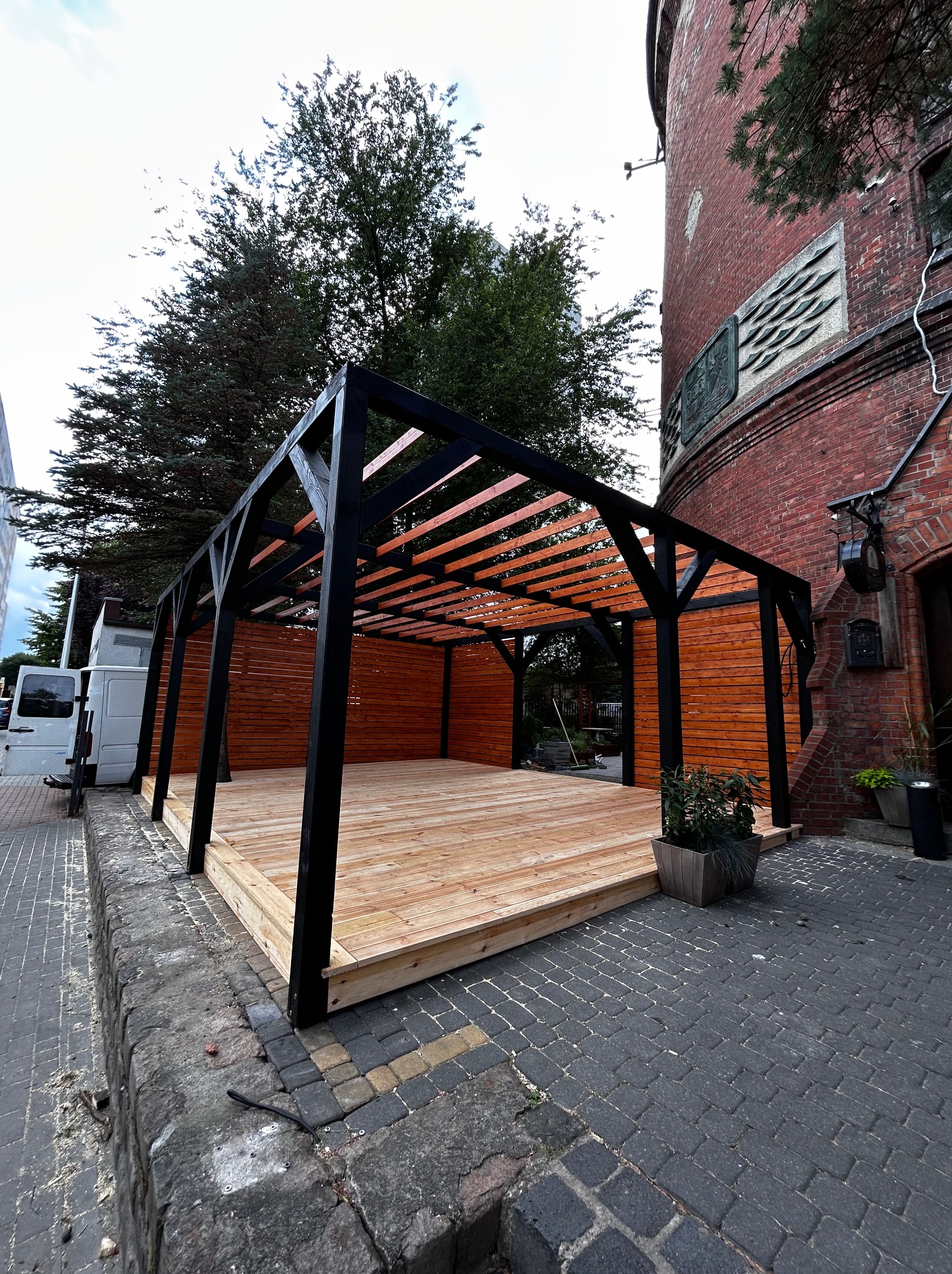Nowoczesna, drewniana pergola z czarną ramą i jasnym drewnem na tle ceglanego budynku i zieleni. Widok z niskiej perspektywy, podkreślający strukturę.