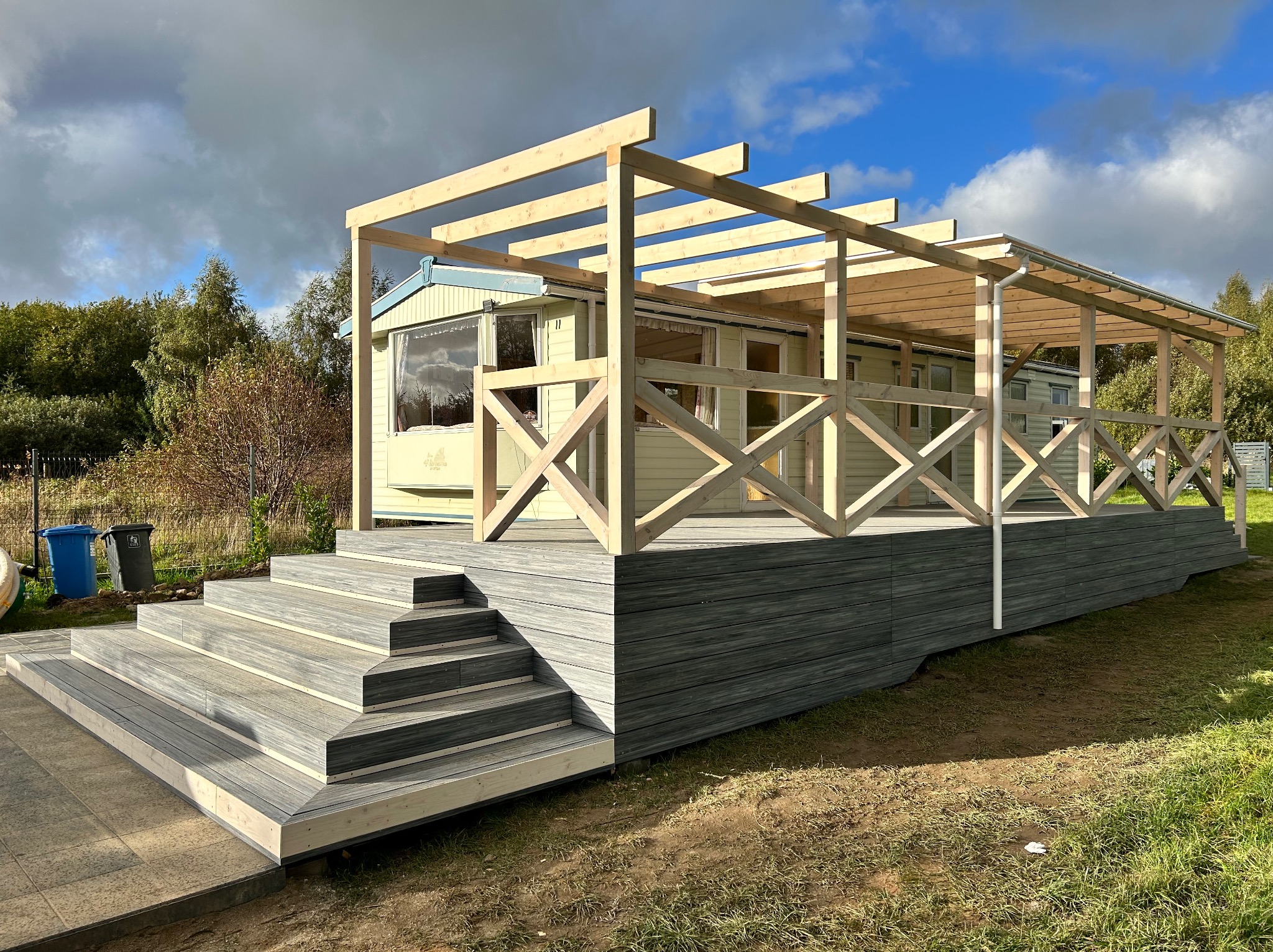 Drewniany taras z balustradą i schodami przed domem, częściowo zadaszony. Konstrukcja w surowym drewnie, widoczne elementy montażowe. Tło stanowią drzewa i błękitne niebo.