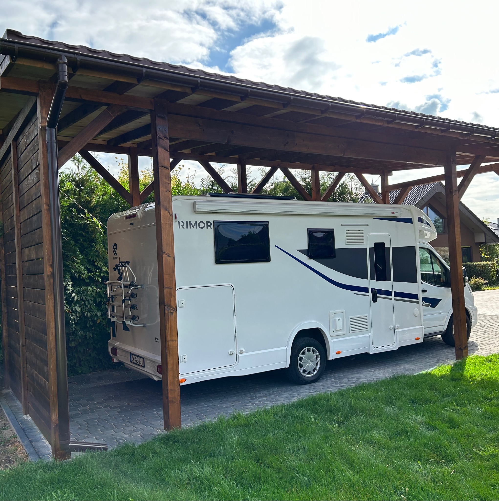 Drewniana wiata garażowa z brązowymi słupami, pod którą zaparkowany jest biały kamper. W tle zielony trawnik i dom.