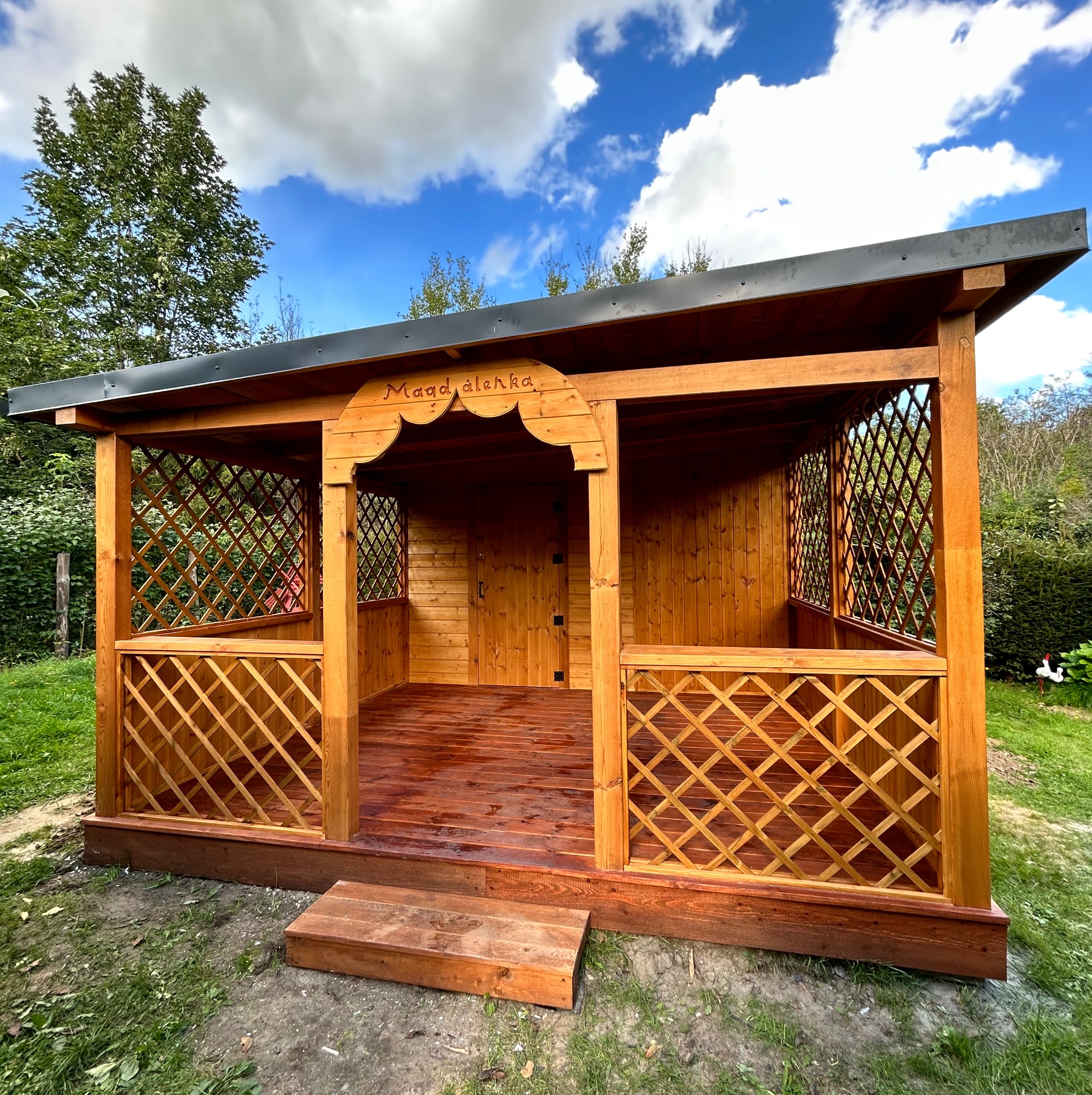 Drewniana altana z kratownicą i napisem 'Maad álenka' pod dachem, na tle błękitnego nieba z chmurami. Widoczny stopień wejściowy i otoczenie z zielenią.