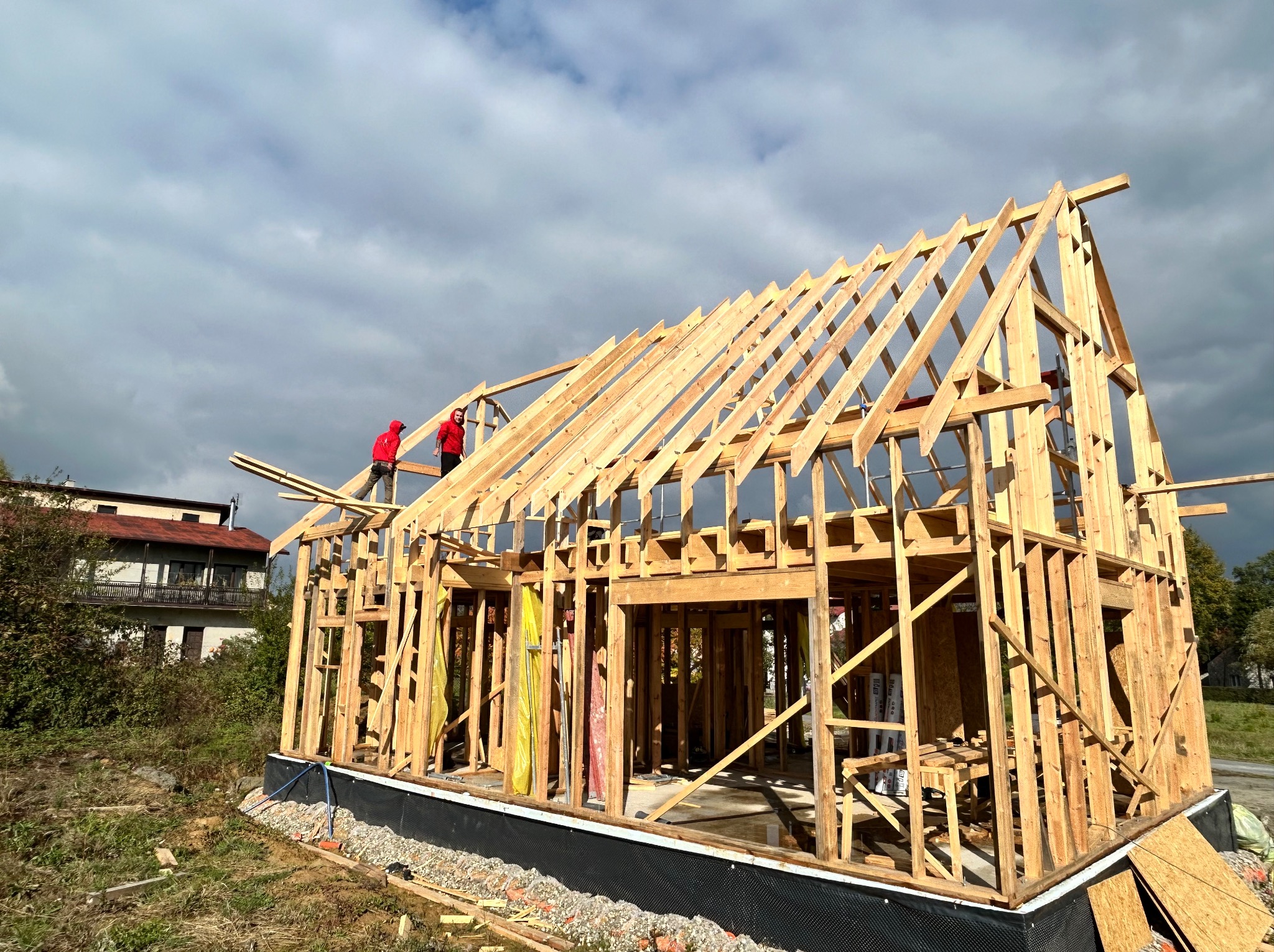 Wznoszenie drewnianej konstrukcji domu szkieletowego. Dwóch pracowników w czerwonych kurtkach montuje elementy dachu. W tle widoczny budynek mieszkalny i pochmurne niebo.