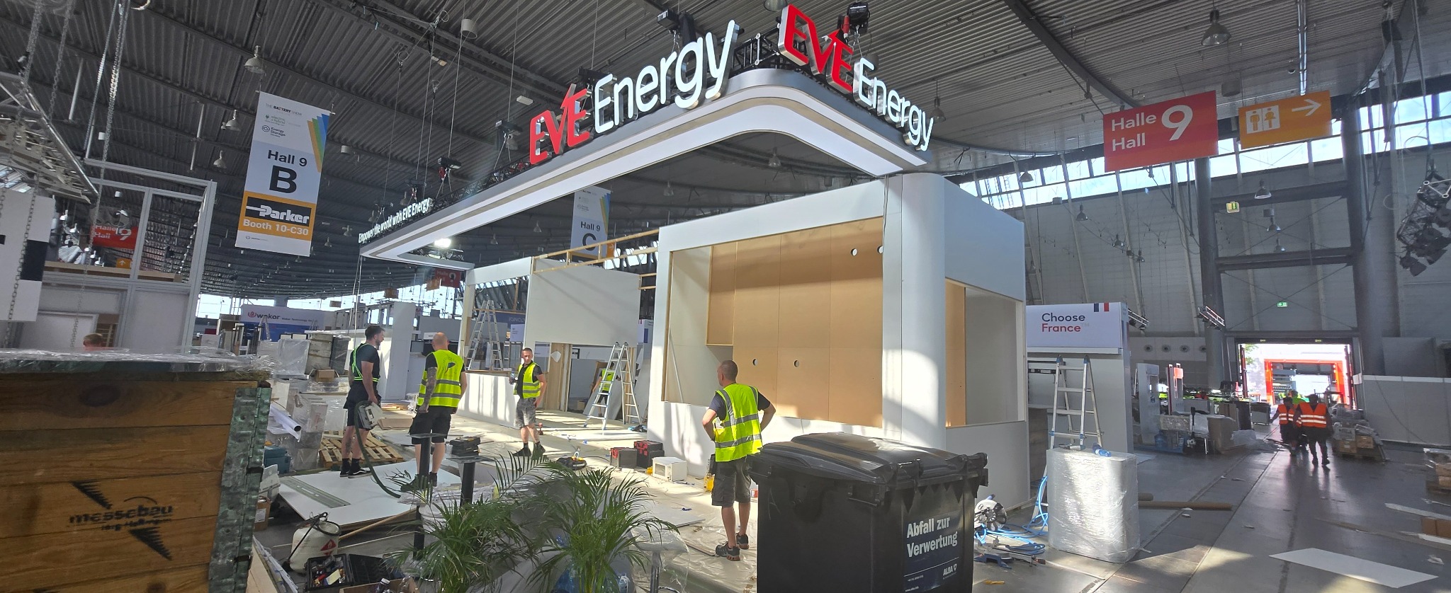 Montaż stoiska targowego EVE Energy w hali: pracownicy w kamizelkach, narzędzia, drabiny, częściowo zamontowane ścianki działowe, rośliny doniczkowe i skrzynie transportowe.