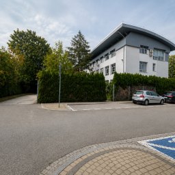 ADWOKAT NYSA - ADWOKAT PAULINA STASIEŁOWICZ - Biały budynek biurowy z charakterystycznym, łukowatym dachem, otoczony zielonymi tujami, z dwoma samochodami na parkingu i niebieskim symbolem dla niepełnosprawnych.