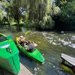 Wypożyczalnia Kajaków Ryś Olsztyn Spływy Łyną - Dwie osoby płynące kajakami po rzece w słoneczny dzień. Jeden kajak zielony na brzegu, drugi na wodzie z parą w kamizelkach ratunkowych.